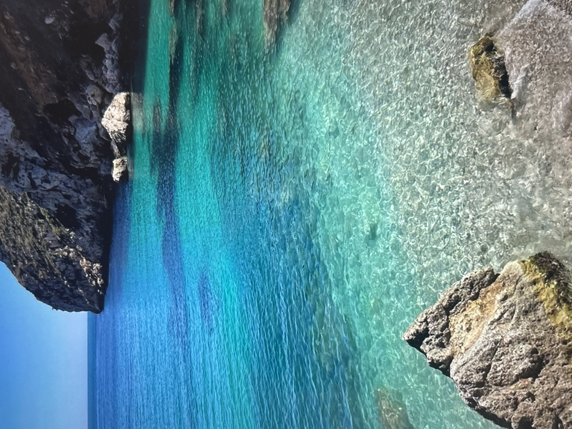 The image presents a vibrant coastal landscape featuring strikingly clear, turquoise-to-blue ocean water bordered by rugged rock formations. The perspective appears to be from above, looking down into a secluded cove or bay.
The central and dominant subject is the water, which showcases a stunning gradient of colors. Towards the left, the water is a deep, rich blue, indicating greater depth, while moving towards the center and right, it becomes a brilliant turquoise and light aqua, suggesting shallower areas where the light-colored seabed is visible. The surface of the water is alive with small ripples and crystalline patterns, reflecting sunlight and highlighting its exceptional clarity.
Dark, imposing rock formations or cliffs frame the water on the left side of the image, extending into the frame's upper edge. Smaller, textured rocks are visible both along the shoreline and partially submerged within the clear water. A prominent, craggy rock formation with a textured, brownish-grey surface juts into the water from the bottom right corner, showing some patches of yellowish-green growth. Another smaller, lighter-colored rock is partially submerged in the very shallow, almost transparent water in the upper right.
The sky, visible in the bottom left corner, is a clear, serene blue, indicating a bright and sunny day. There are no people, animals, or any specific activities taking place; the scene is purely a natural landscape. The overall impression is one of untouched natural beauty, tranquility, and remarkable water clarity. No visible text is present in the image.
