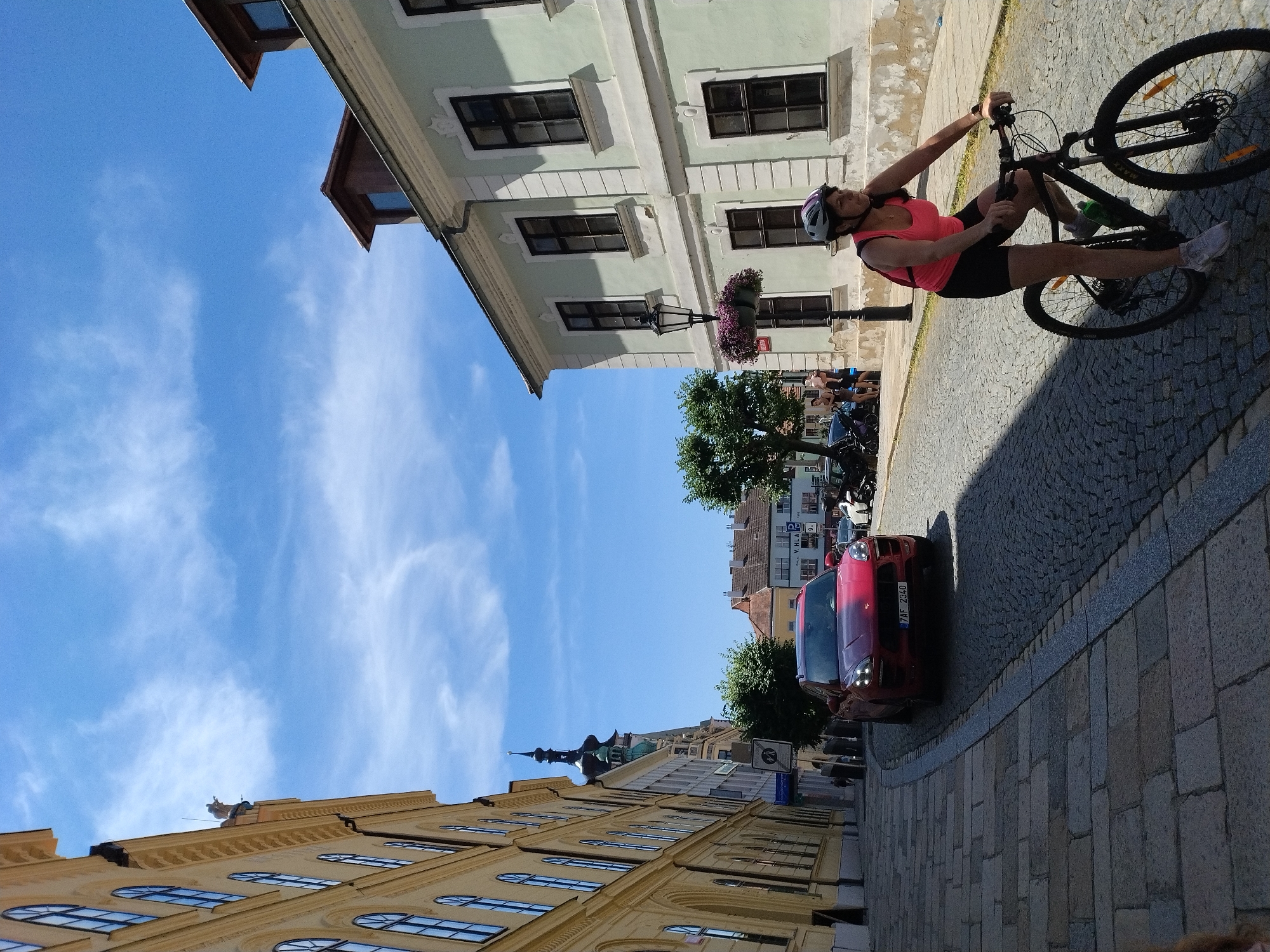 In Písek, Czechia, a woman wearing a pink tank top, black shorts, and a white helmet with purple accents rides a black bicycle with a green water bottle attached to the frame. She is on a cobblestone street with a red car parked to her left. The car has a license plate that reads "7AF 2340". To the left of the street, a tall yellow building with arched windows lines the street. To the right of the street, a light green building with dark windows stands. In the distance, a church tower with a green dome can be seen. The sky is blue with scattered white clouds.