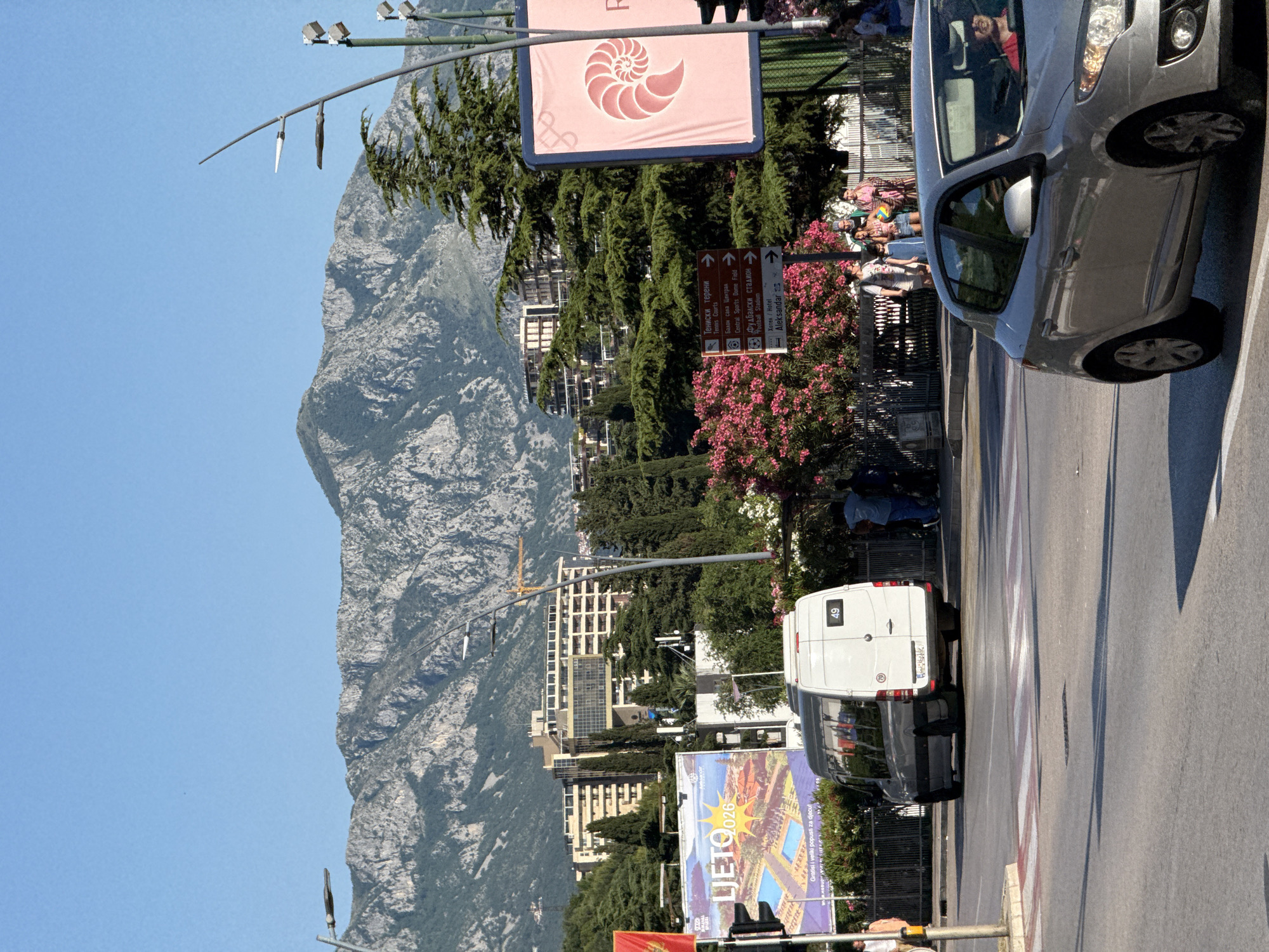 The image depicts a vibrant street scene in Budva, Montenegro, under a clear blue sky. Dominating the background are rugged, rocky mountains, with several multi-story buildings, including a large hotel-like structure, nestled amongst dense green vegetation, including conifers and flowering pink oleander bushes.
In the foreground, a silver Renault Megane hatchback is turning right, with its driver visible. A white Mercedes-Benz Sprinter van, marked with "49" on its rear and a partially visible Montenegrin license plate ("BP"), is stopped on the road near a zebra crossing, facing away from the viewer.
On the left, a large billboard with "LJETO 026" and an image of a resort pool is prominent, featuring text in Cyrillic. Adjacent to it, a red and yellow Montenegrin flag is partially visible. In the center, a brown directional sign post lists locations in both Serbian Cyrillic and English: "Тениски терени / Tennis Courts," "Базен Сала Центар / Central Sports Hall," "Фудбалски стадион / Football Stadium," and "Хотел Александар / Hotel Aleksandar." To the right, a large pink billboard displays a stylized spiral shell logo.
Several pedestrians are visible on the sidewalks, including a group of adults and a child holding a colorful beach ball near the pink flowering bushes on the right. Streetlights and power lines are present overhead. The scene is illuminated by bright sunlight, casting distinct shadows.