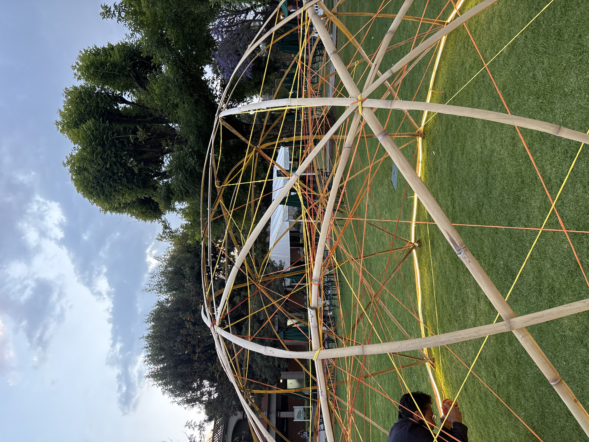 This image depicts an outdoor area in San Andrés Cholula, Mexico, observed during twilight. A large, partially constructed dome structure occupies the central and foreground. The dome is composed of light-colored bamboo poles forming its skeletal framework, interlaced with a dense network of thin orange and yellow synthetic cords. A circular ring of yellow LED strip lighting illuminates the perimeter of the dome's base, which rests on vibrant green, manicured grass.
In the lower-left foreground, a solitary individual, visible from the mid-torso upwards, is actively engaged within the dome. Dressed in a dark long-sleeved shirt, the person is bent over, manipulating an orange cord with both hands, indicating an ongoing assembly or adjustment process.
The background features a building with a red-tiled roof and a light-colored facade on the left. To the right, a white event tent is visible, accompanied by several green patio umbrellas and dark outdoor tables with chairs arranged on the grass, suggesting a temporary event setup or a dining area. Numerous mature trees with extensive green canopies, one displaying subtle purple blossoms, line the perimeter of the area against a sky with scattered grey and white clouds.
