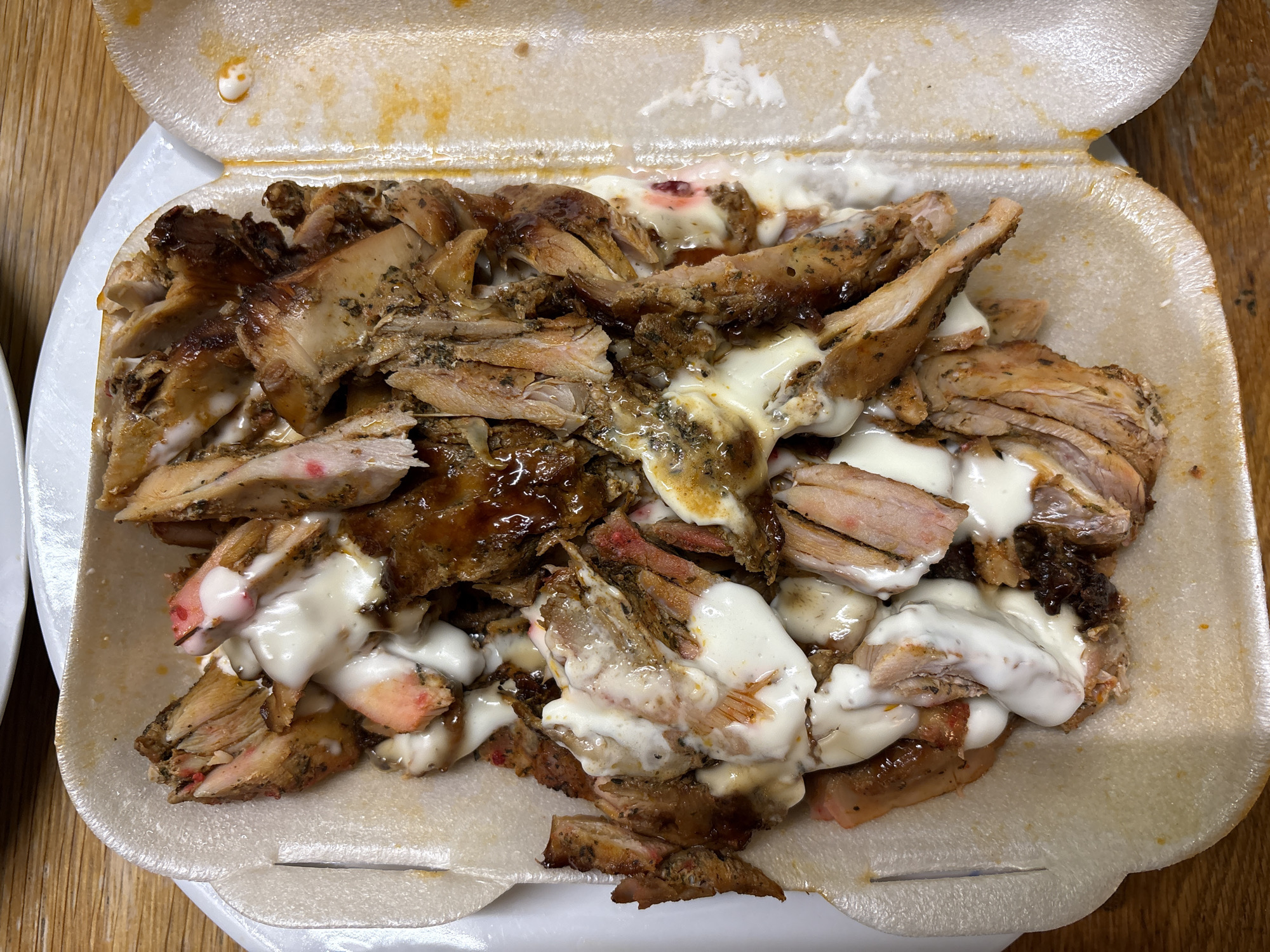 The image shows a close-up of sliced meat with white sauce in a white styrofoam container. The meat appears to be a mix of light and dark pieces, some glazed with a darker sauce. The container is rectangular with rounded corners and is sitting on a wooden surface. The location is Oswestry, United Kingdom. There are no people visible.