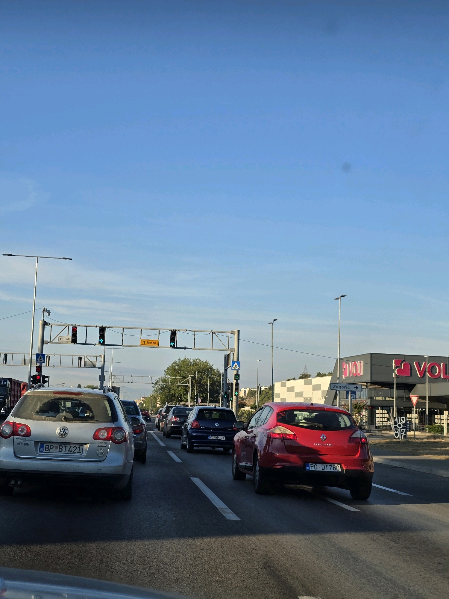The image captures a traffic scene in Podgorica, Montenegro, under a clear blue sky. The foreground shows the rear of a silver Volkswagen Passat wagon, license plate BP BT 421, in heavy traffic. To its right, a red Renault Megane hatchback, license plate PG DT780, is also in traffic. Further ahead, several other cars are visible, including a dark blue Volkswagen Golf.
In the background, traffic lights are positioned overhead, with some showing red and others green. A sign indicating "Tranzit" (Transit) is also visible. To the right, a modern building with prominent "VOLI" signage stands, likely a supermarket or retail complex. A directional sign points towards "Zagorje". Streetlights and utility poles are present along the road. The environment suggests a well-developed urban area with active commerce and infrastructure. No individuals are clearly discernible in the image.