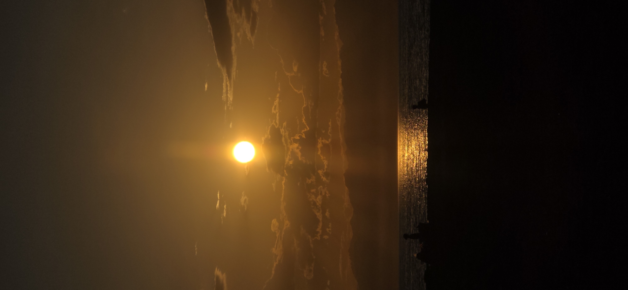 A vertical, full-frame shot captures a sunset over the sea at Boz Beshik, Kyrgyzstan. The sun, a bright, orb-like sphere with a luminous halo, hangs centrally in the sky, casting a golden path of light across the rippling water. Clouds, tinged with orange and gold from the setting sun, are scattered across the sky, with some appearing darker and more solid. In the foreground, silhouetted against the bright water and sky, are the dark shapes of people and possibly beach furniture on a sandy shore. The overall impression is one of tranquility and natural beauty.