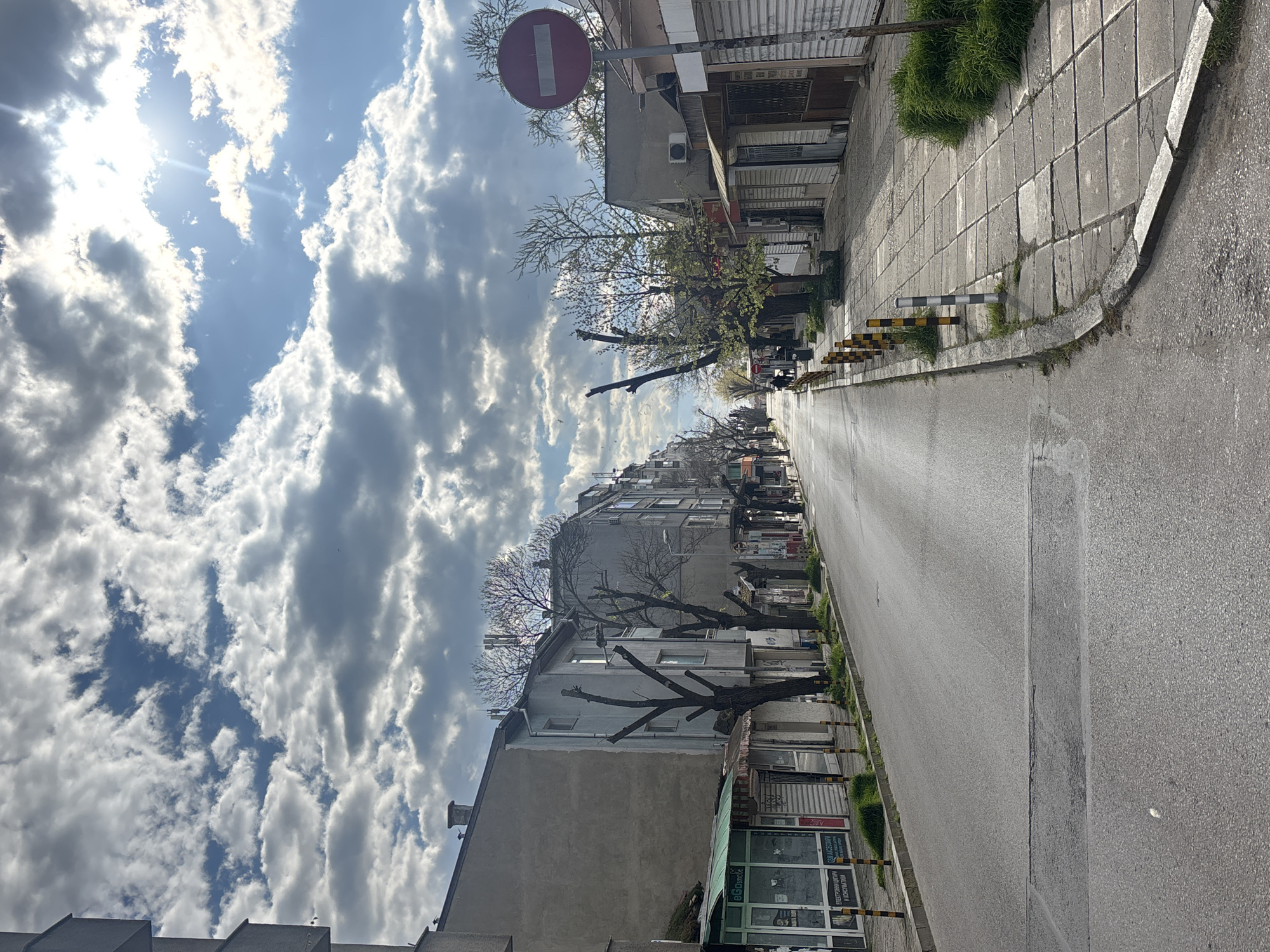 The image depicts a street view in Varna, Bulgaria. The street is lined with buildings of varying heights and architectural styles, most of which appear to be residential or mixed-use, with shops at the ground level. Bare trees with pruned branches are visible along the street. A "no entry" road sign is posted at the beginning of the street. The street is paved and appears wet or recently cleaned. The sky is cloudy with patches of blue. There are no people clearly visible in the image. The focus is on the urban environment and the weather conditions.