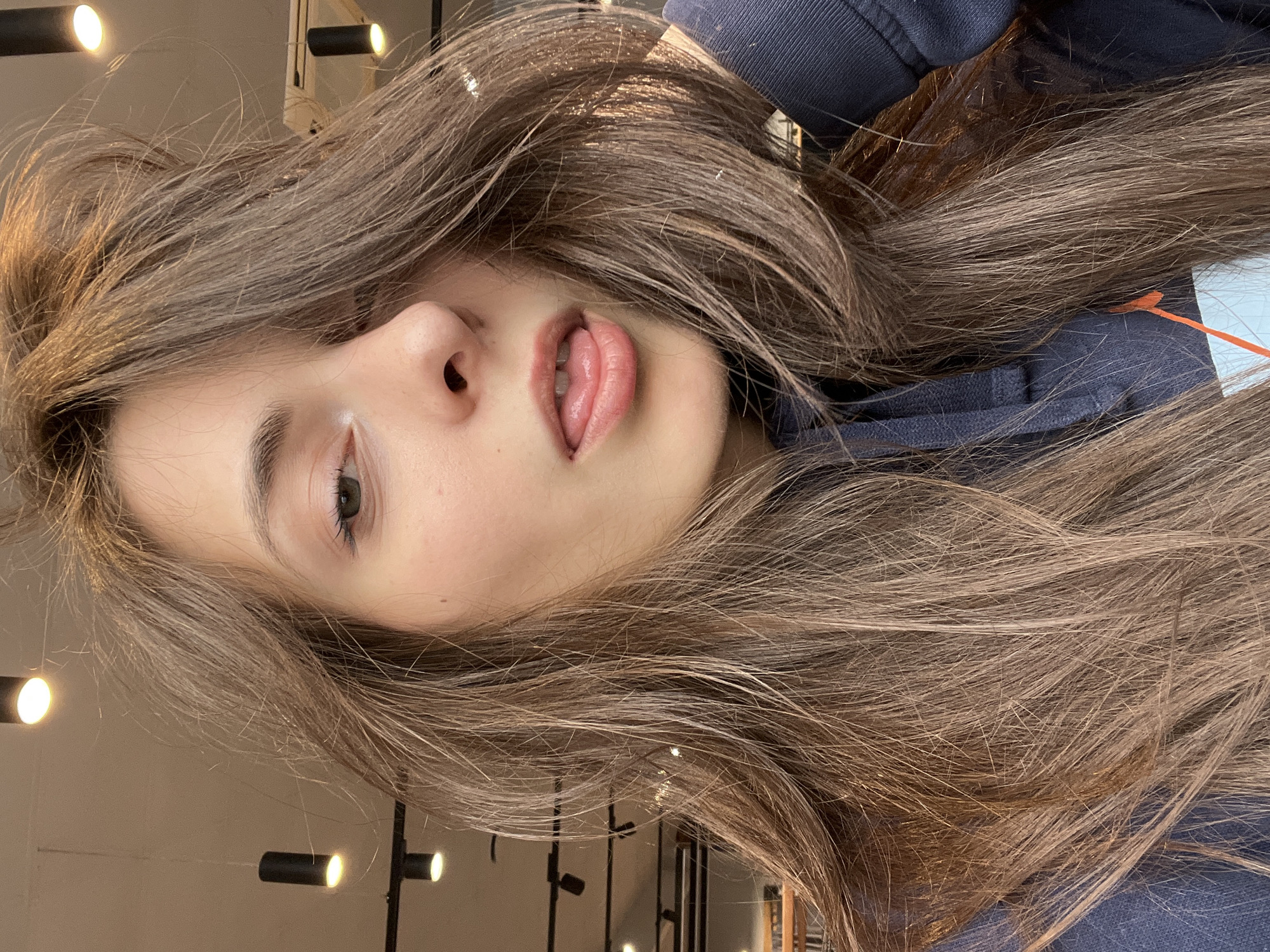 A medium close-up portrait captures a young woman, with the image rotated ninety degrees counter-clockwise. She appears to be lying on her side, looking towards the viewer with a casual and playful expression. Her long, flowing hair is a warm light brown with subtle auburn highlights, cascading around her head and shoulders. The hair appears thick, slightly wavy, and somewhat artfully disheveled.
Her face is the central focus, displaying fair and smooth skin with a natural sheen on her nose and cheekbones. Her eyes, appearing to be a light color, are partially open and engaging with the camera. Her mouth is slightly open, with her tongue playfully peeking out between her lips. She has well-defined eyebrows and visible eyelashes.
She is wearing a dark navy blue hooded garment, possibly a hoodie or jacket, parts of which are visible around her neck and shoulders. A notable detail is a small white tag with a distinct orange arrow-like graphic, visible near her right shoulder peeking through her hair. A hint of an orange cord is also visible in this area, potentially from headphones or a drawstring.
The background is softly blurred, indicating an indoor setting. Several modern light fixtures are visible, appearing as small, cylindrical spotlights, some track-mounted and others as pendants. These lights emit a warm, golden glow. The overall lighting is bright and soft, highlighting the subject's features. The scene suggests a relaxed moment in a contemporary interior, such as a cafe, office, or home, located in Krasnoyarsk, Russia. There is no legible text visible beyond the graphic on the small clothing tag.