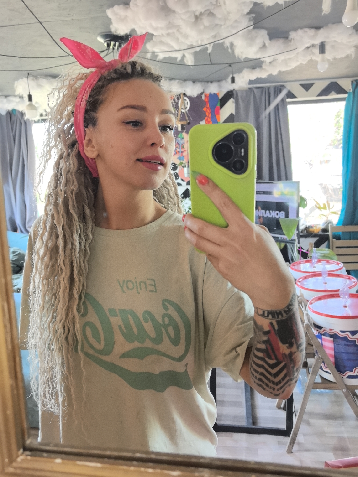A mirror selfie shows a woman with long, blonde dreadlocks and a pink headband with bunny ears. She is holding a green smartphone in her right hand, obscuring part of her face. She wears a light green t-shirt with a large, dark green "Enjoy" graphic. A tattoo is visible on her right forearm. The background includes a room with decorative elements such as artificial clouds suspended from the ceiling, patterned curtains, a window, and what appears to be a television screen showing "BOKUHM." Several buckets with red lids are stacked on a folding table in the foreground. The exact location within the city of Noviy, Russia, cannot be determined from the image.