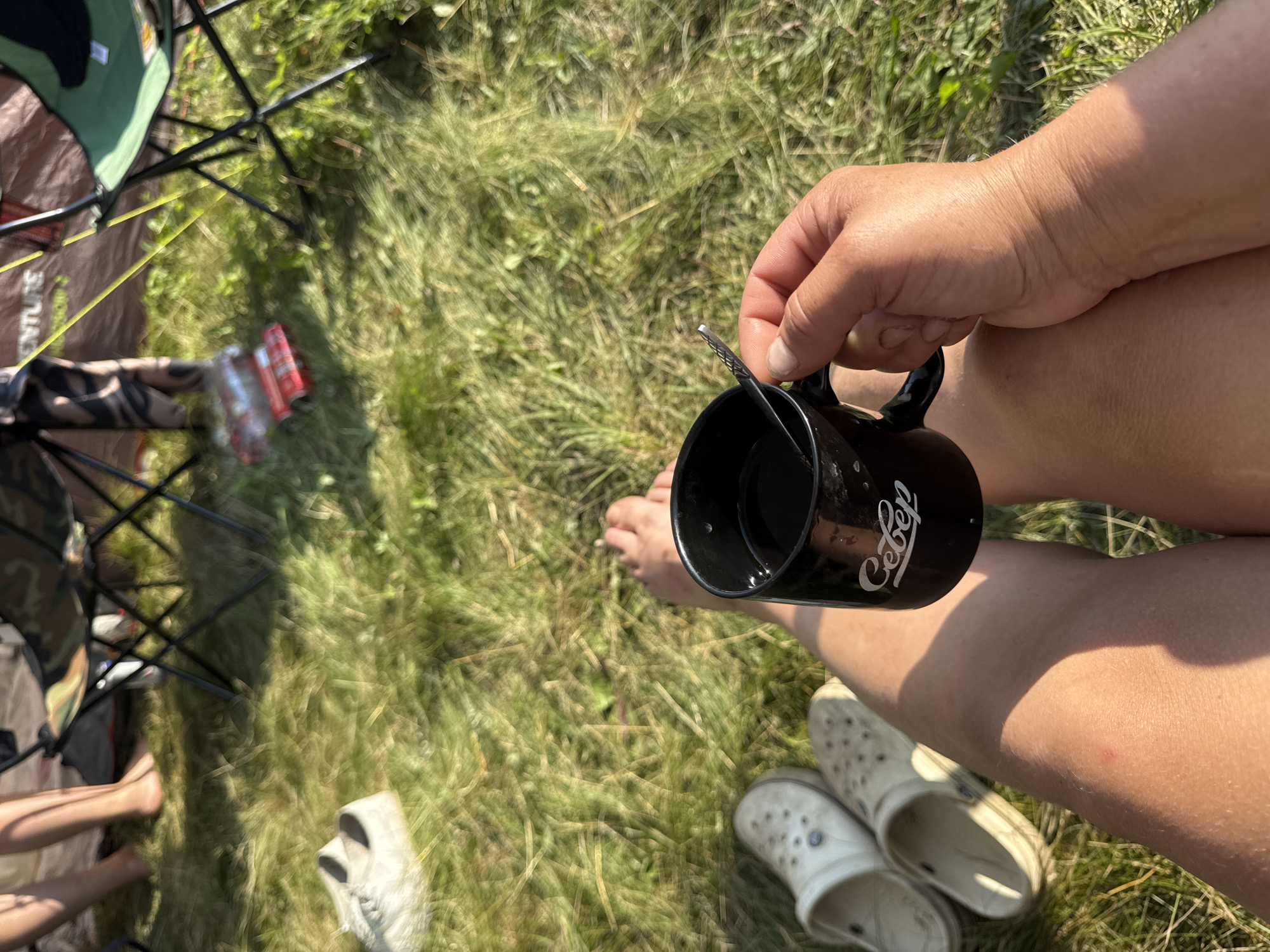A first-person perspective shot captures a person's legs, a black mug, and a hand holding the mug, set against a backdrop of lush green grass and camping equipment. The person is sitting with their legs crossed, one foot bare and the other resting on a white Croc shoe. Another white Croc shoe is visible next to it. In the background, several camping chairs are set up, one in camouflage pattern and another in green. A tent, partially visible, has the word "ADVENTURE" printed on it. Scattered on the grass are packets of what appear to be snacks or cigarettes. The scene suggests a relaxed outdoor setting, possibly a campsite.