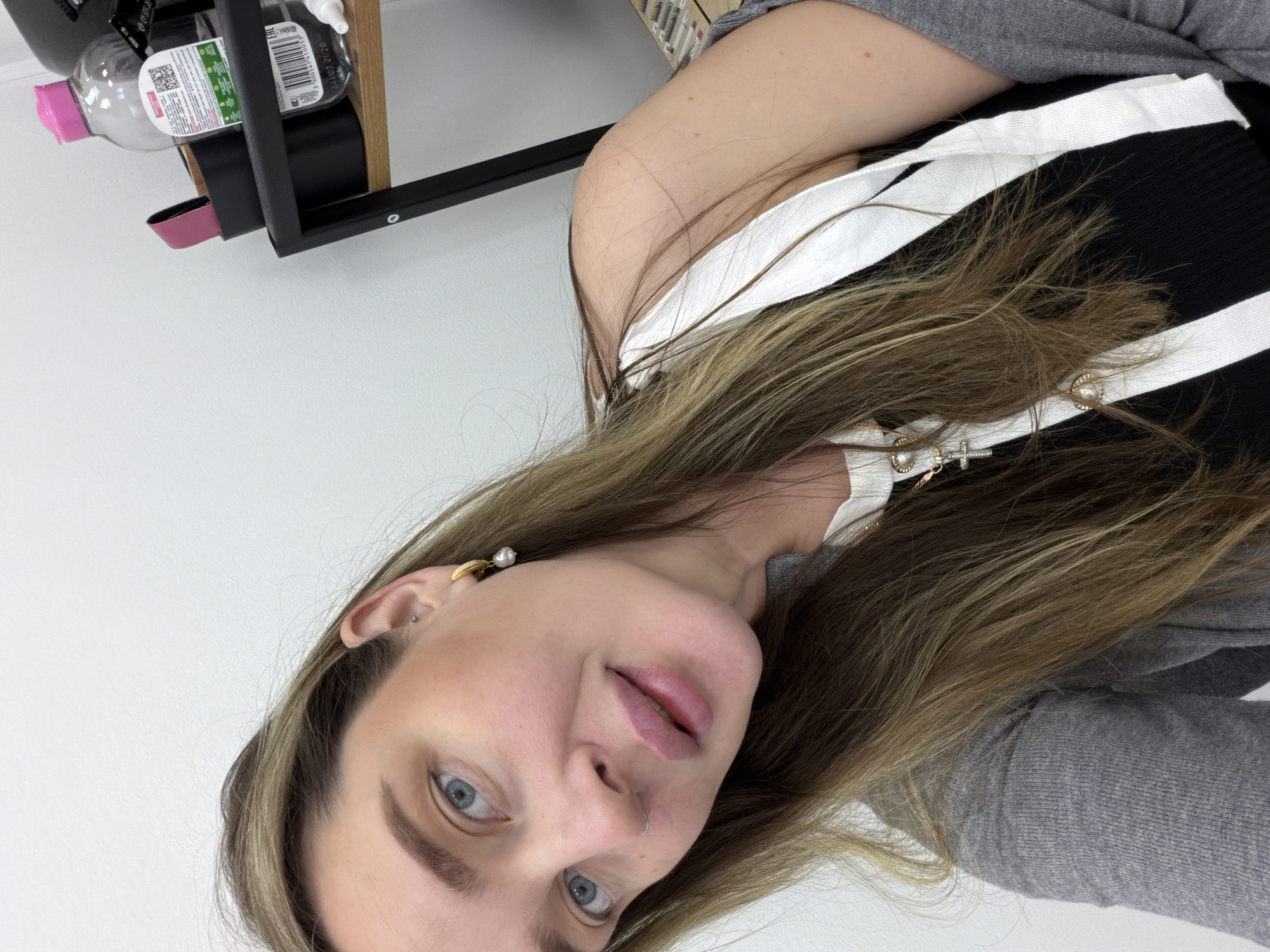 A young woman with long, light brown hair and blue eyes is looking directly at the camera in a close-up, slightly angled shot. She has fair skin, visible freckles, and a neutral expression with slightly parted lips. She wears a nose ring (septum piercing) and an ear piercing, along with a gold earring featuring a pearl on one ear. Her eyebrows are well-defined.
She is dressed in a black and white striped top layered under a grey long-sleeved garment, possibly a cardigan or sweater. Several necklaces are visible, including one with a small gold cross pendant and another with a circular gold charm. Her hair is worn down, partially covering her shoulders and chest.
The background is a plain white wall. To the top left, a dark-colored shelf or frame is visible, holding several items. Among these are clear plastic bottles, one with a pink cap. Partial text and a barcode are visible on one of the bottles. Visible text includes "EAC", a QR code, and numbers like "38340", "5 000104 00029", and "01.29". The setting appears to be indoors under artificial lighting. Based on the provided context, the image location is Moscow, Russia.