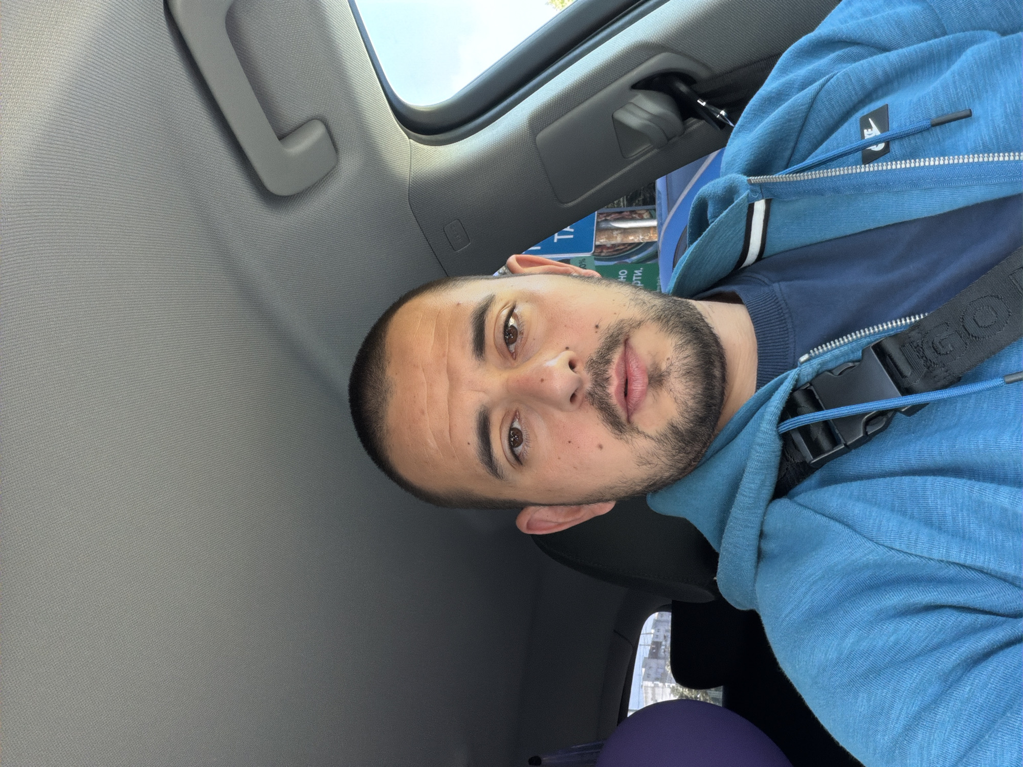 A male individual, appearing to be in his 30s, with a closely shaved head and a short beard, is seated in the driver's seat of a car, facing forward and looking directly at the camera. He wears a blue zippered hoodie with a white Nike logo on the left chest, layered over a darker blue long-sleeved shirt. A black strap, identifiable by "IGOL" lettering, crosses his chest from his right shoulder to his left hip. The car's interior features a light gray fabric headliner, a gray passenger-side grab handle, and a visible "AIRBAG" label on the passenger A-pillar. A black seatbelt is buckled across the individual. In the rear of the car on the left side, a partially visible purple balloon is present. Outside the car, through the passenger window, a clear blue sky and bright sunlight are visible. A street sign with partial Cyrillic text, specifically "ТА" and "НО РТИ", is visible, consistent with the Bulgarian language. Urban buildings and a blue vehicle or structure are present in the background, placing the scene in a city environment in Burgas, Bulgaria.