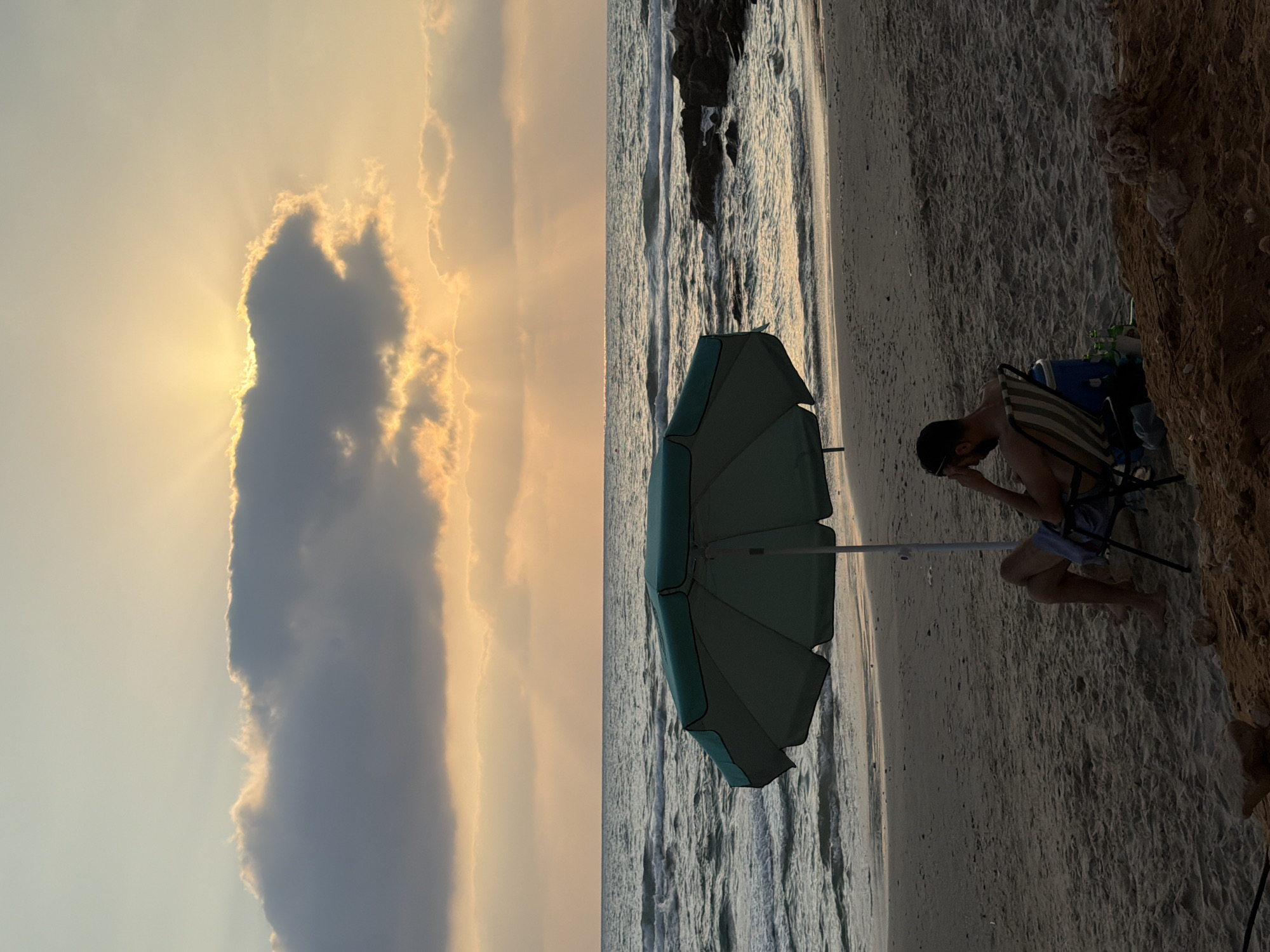 The image depicts a serene sunset scene on the beach of Hadera, Israel. A lone man sits in a striped beach chair under a green umbrella, his hand resting on his head, gazing towards the sea. The ocean waves gently lap the sandy shore, and the sky is painted with hues of orange and yellow as the sun sets behind a large cloud, casting ethereal rays of light. Rocks are visible in the water near the shore. Several bottles and a blue cooler are placed beside the man's chair. The sand is textured with footprints and small pebbles.