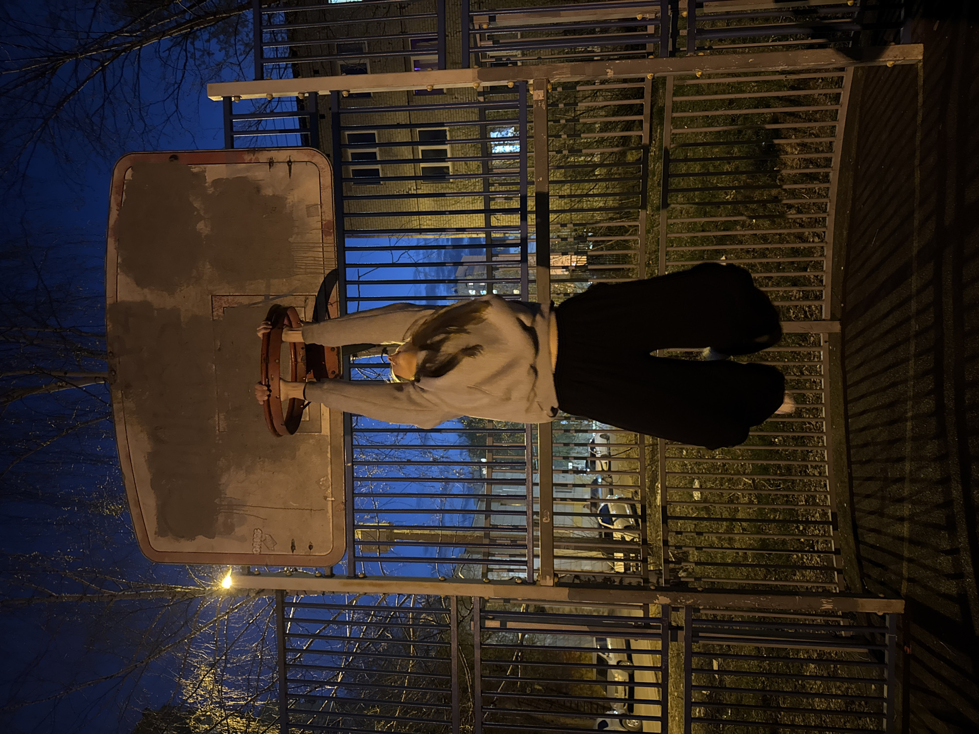 The image shows a person hanging on a basketball hoop at night. The person is wearing a gray sweatshirt and black pants. The basketball hoop is old and rusty, with a concrete backboard. Behind the basketball hoop is a metal fence and a building with lit windows. Trees with bare branches are visible in the background. The location is likely in Perm, Russia.