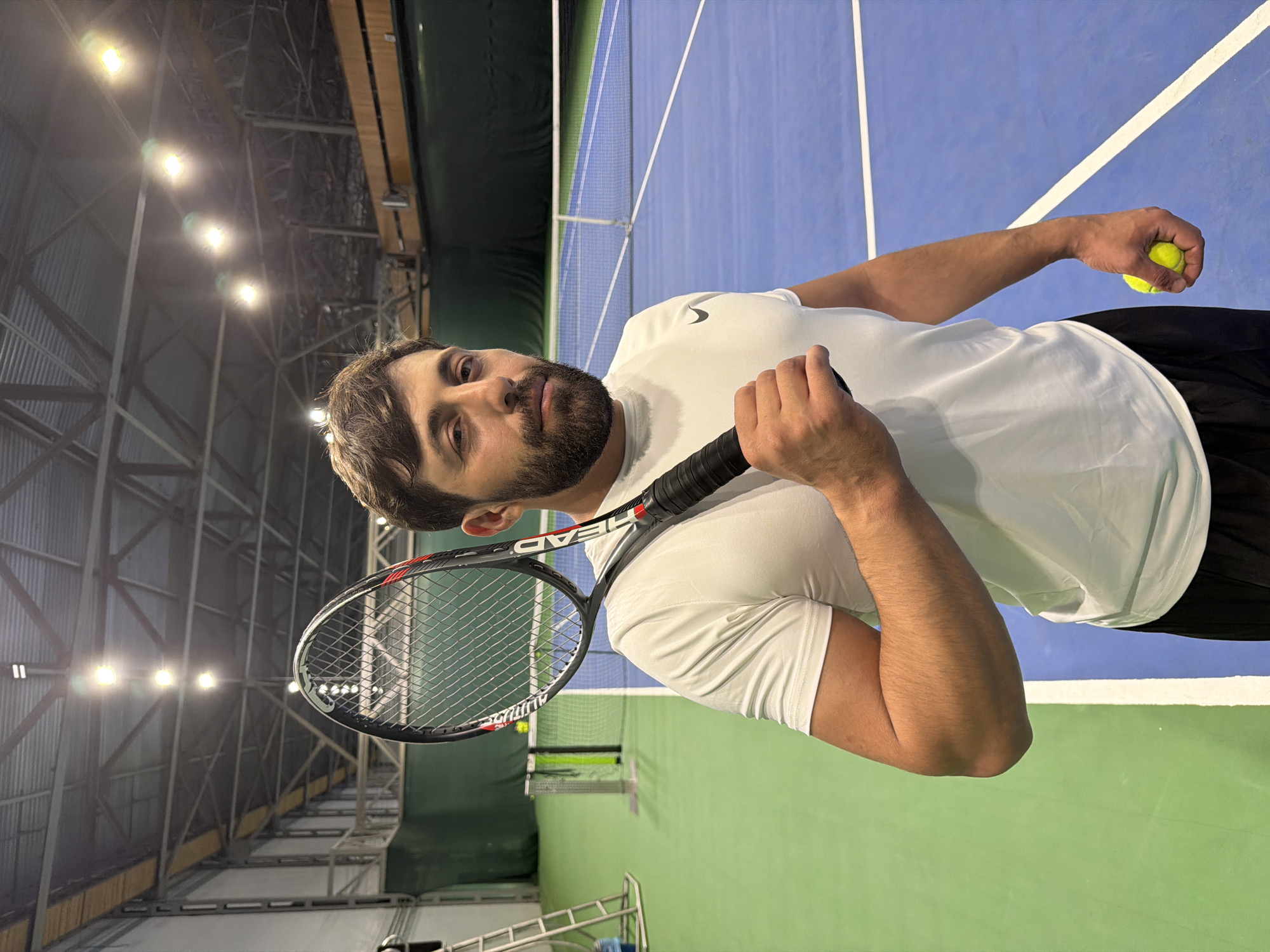 A man with a beard, wearing a white t-shirt and black shorts, stands on an indoor tennis court holding a tennis ball in his right hand and a tennis racket in his left. The court has blue and green sections, with white lines marking boundaries. A net is visible in the background separating the court. Overhead, there are industrial-style lights illuminating the court. The location is likely inside a sports complex within the city of Krasnoyarsk, Russia.