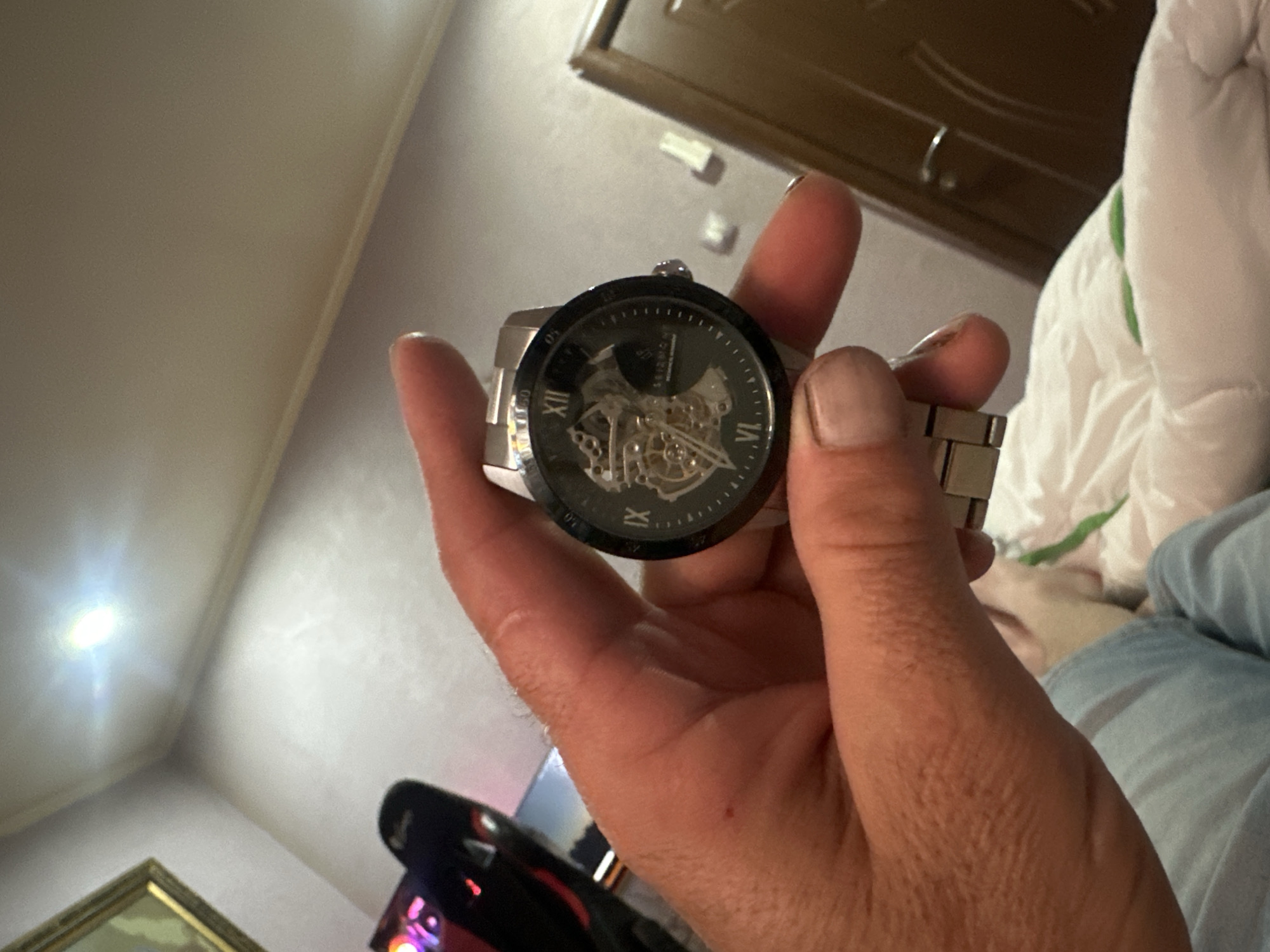 The image shows a close-up of a person's hand holding a silver-toned skeleton watch with a black dial. The watch has Roman numerals for hour markers and a visible inner mechanism. The background is blurred but appears to be an indoor setting with a ceiling light and a partially visible door. There are no recognizable landmarks or features to indicate the location in Cherven Breg, Bulgaria.