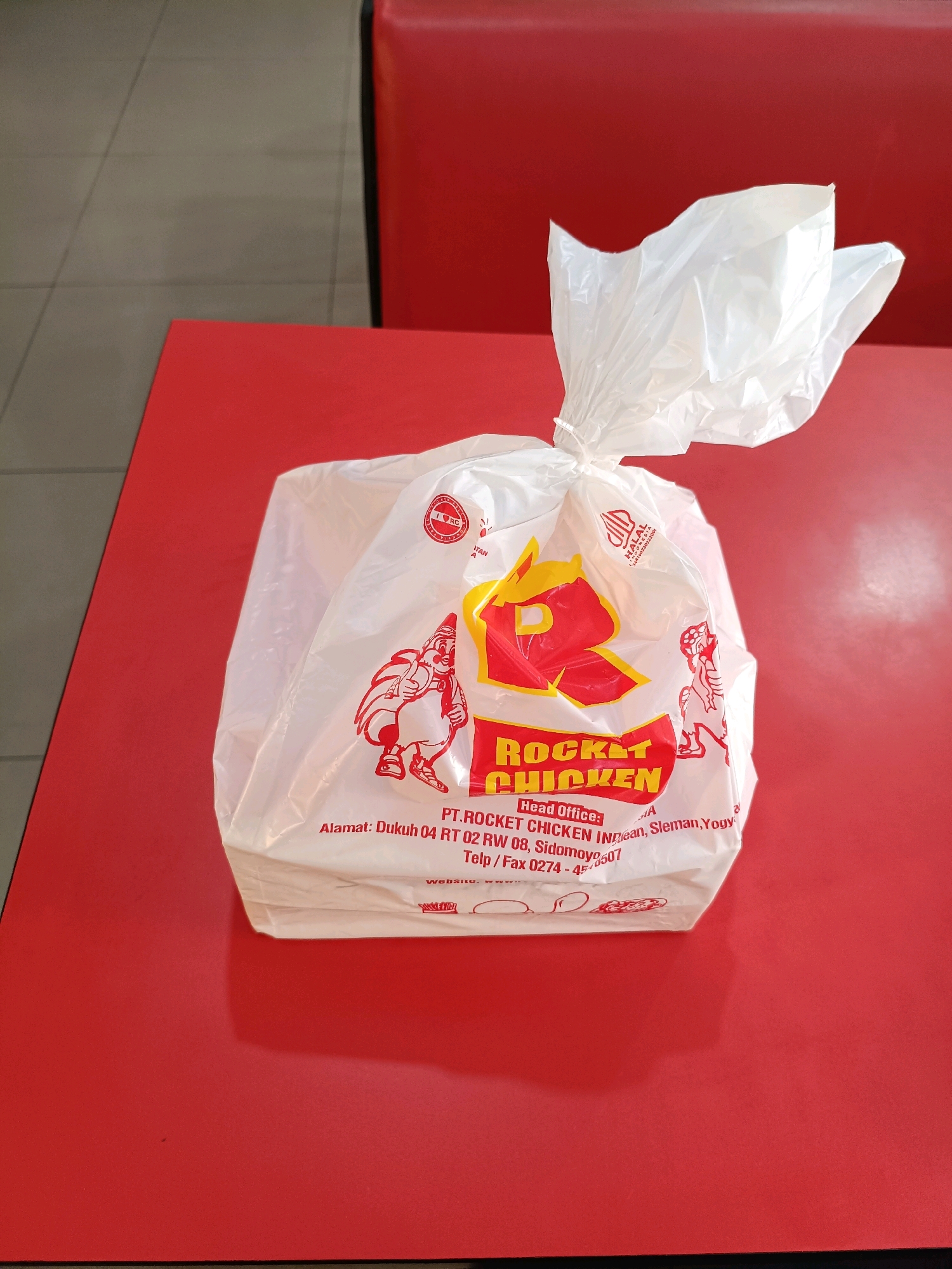 A white plastic bag with red lettering and graphics sits on a red table. The bag is tied at the top with a white plastic band. The lettering on the bag reads "ROCKET CHICKEN." Below this, the text "Head Office: PT. ROCKET CHICKEN INDONESIA" is visible, followed by an address that includes "Dukuh 04 RT 02 RW 08, Sidomoyo, Ngemplak, Sleman, Yogyakarta." A phone/fax number is also printed on the bag. The bag appears to contain food. The table surface is solid red. In the background, a red booth-style seat is visible. The floor appears to be tiled in a light gray color. The image is taken from a slightly elevated, eye-level perspective, looking down at the bag.