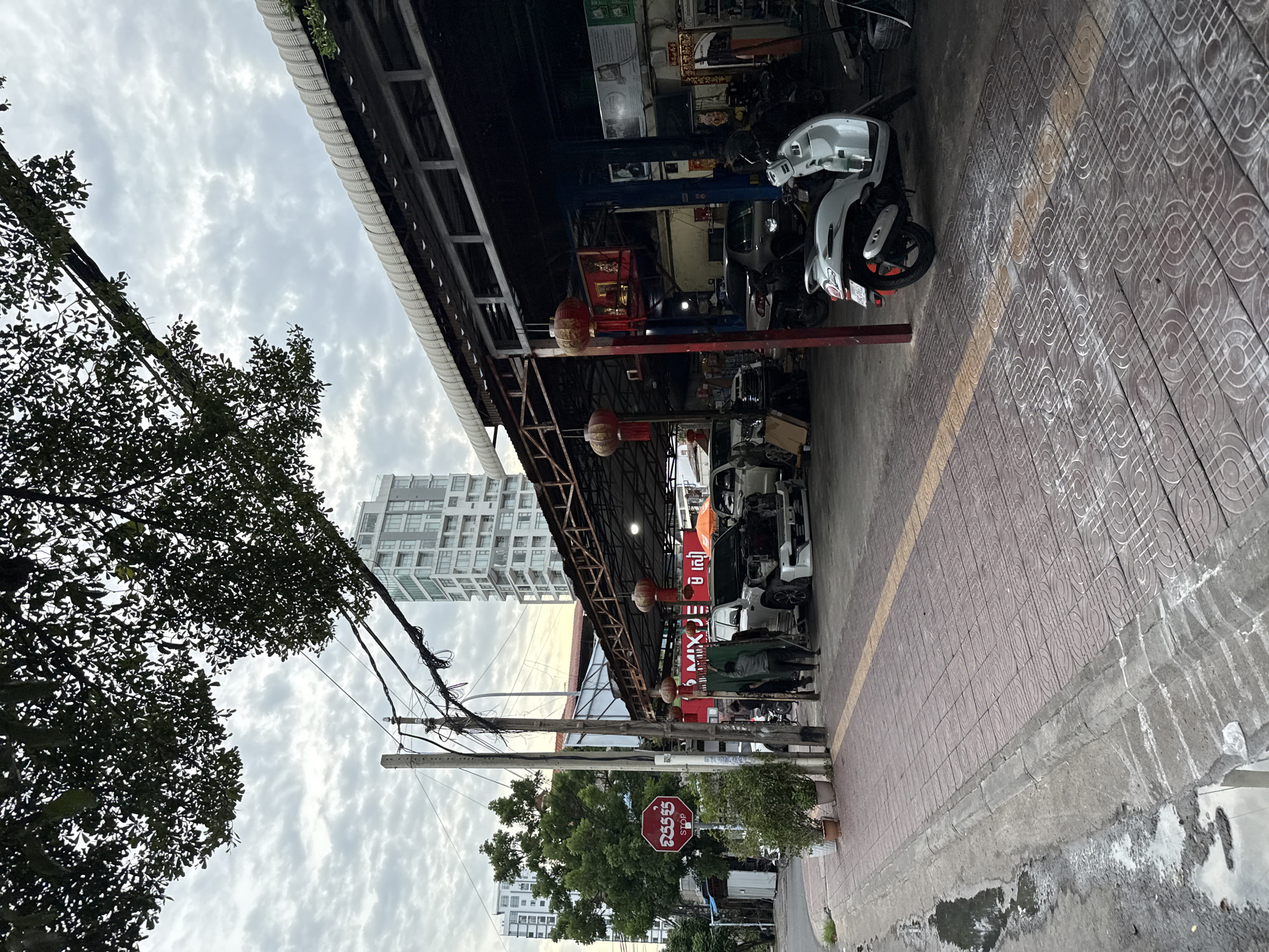 The image captures an overcast day in Phnom Penh, Cambodia, specifically in the Khan Daun Penh district. A car repair shop is visible, with several vehicles, including a white SUV and a white scooter, present. One person, dressed in dark clothing, is standing near the SUV. Red lanterns hang from the shop's awning, adding a decorative element. Electrical poles with numerous wires stand on the sidewalk, which is paved with patterned tiles. A red stop sign with Khmer script is also visible on the sidewalk, alongside potted plants. In the background, modern high-rise buildings can be seen, contrasting with the workshop and surrounding urban environment. Puddles of water are present on the street next to the sidewalk.