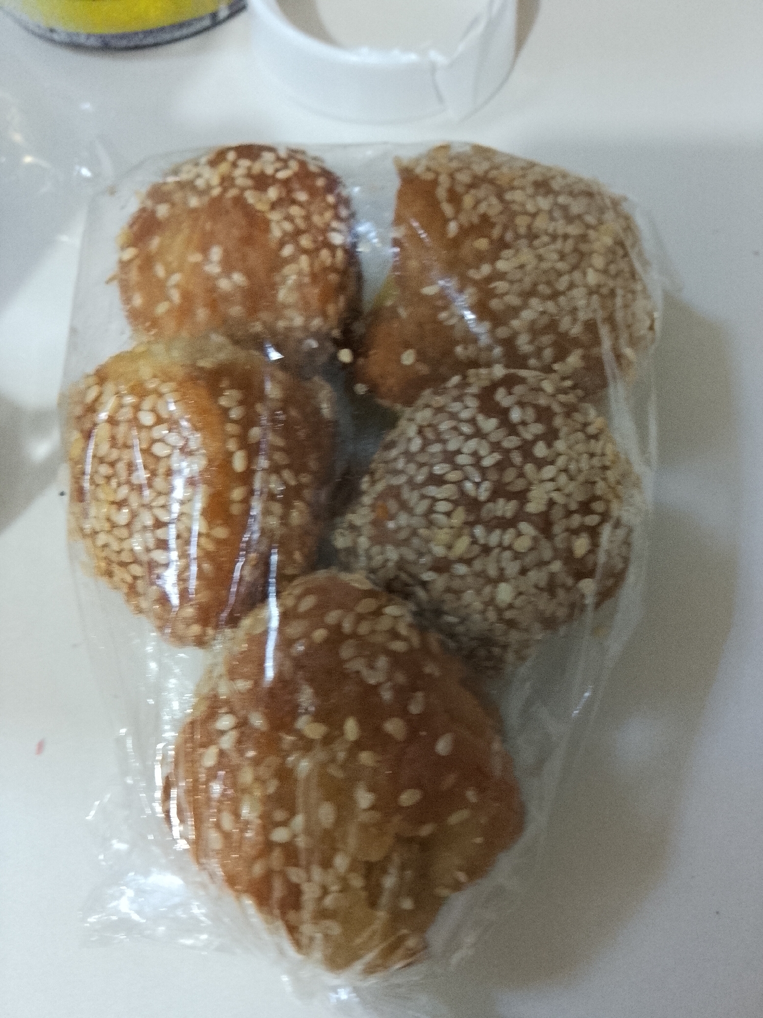 Five individual, irregularly shaped, golden-brown pastries are depicted in the foreground, each heavily coated with white sesame seeds and enclosed in clear plastic wrapping. The pastries vary slightly in form, appearing somewhat rounded or ovular, with a visible texture from the baked surface and the dense sesame seed application. They are arranged in a cluster within the frame. The background consists of a light-toned, uniform surface, likely a tabletop or countertop. Blurred objects are present in the upper left and upper center of the image; a yellow object (possibly a container lid) is partially visible on the left, and a white object (potentially another container or dish) is visible centrally. The lighting is ambient, producing slight reflections on the plastic packaging. The scene is located in Davao City, Philippines.