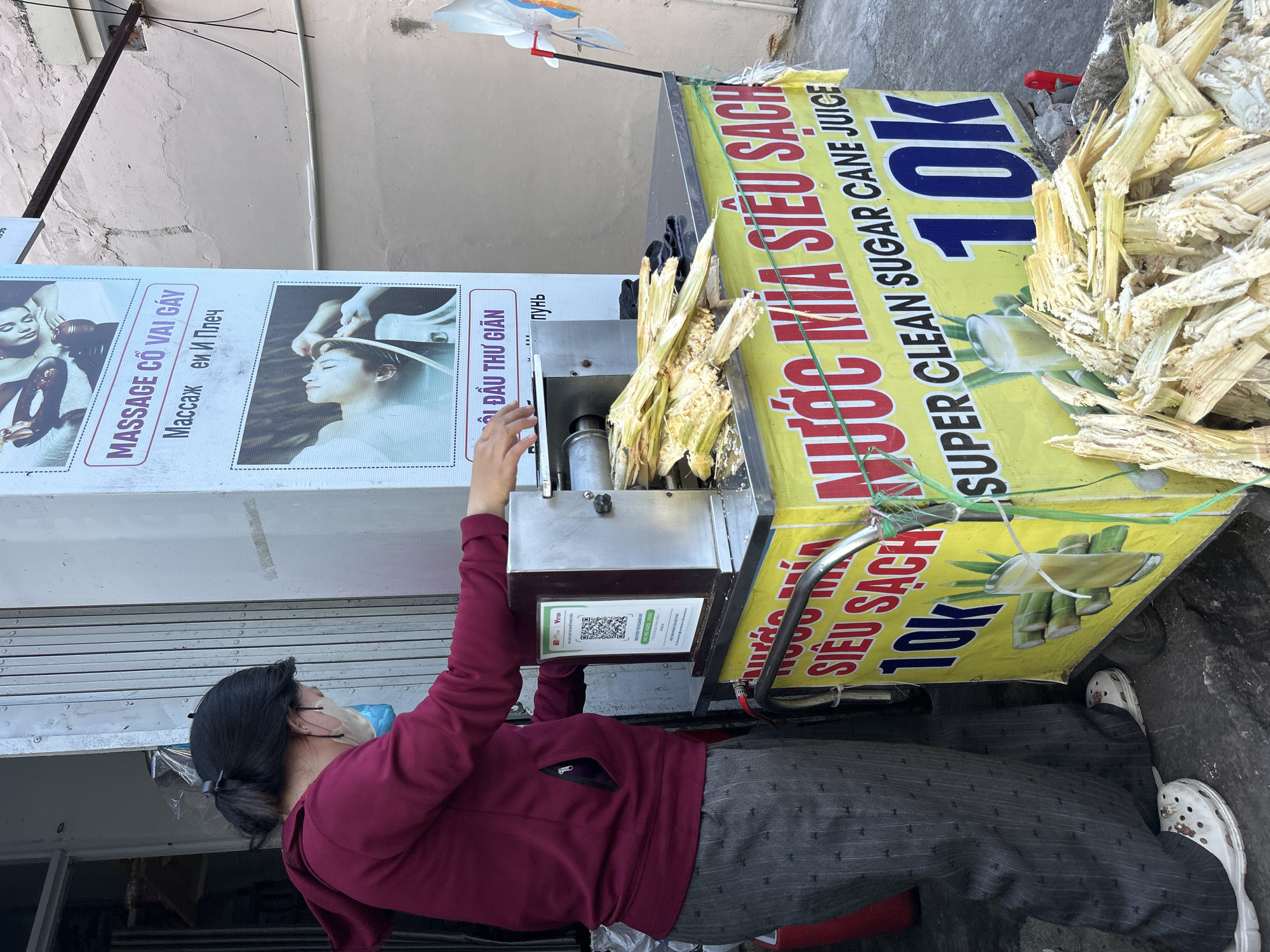 Here is a description of the image:
The image shows a woman wearing a mask and a red sweater, operating a sugarcane juice cart on a street in Bắc Nha Trang, Vietnam. She is feeding sugar cane into a machine. The cart features a yellow and red advertisement for "NƯỚC MÍA SIÊU SẠCH" (Super Clean Sugarcane Juice) with the price "10K". The cart is full of peeled sugarcane. Behind the cart is a sign advertising massage services in Vietnamese, Russian, and English.