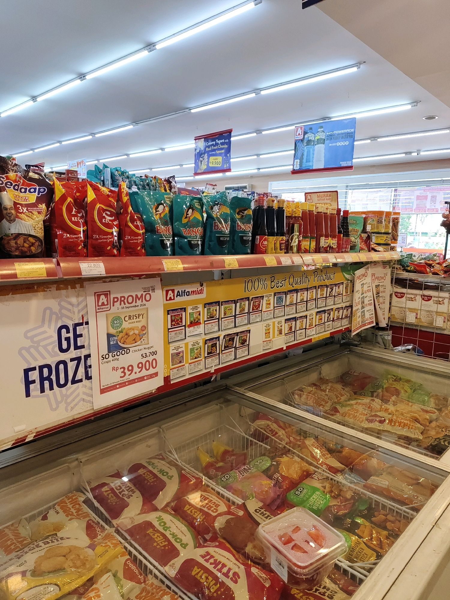 The image depicts the interior of a supermarket aisle in Probolinggo, Indonesia, specifically an Alfamidi store. The aisle is lined with shelves stocked with various food products. Prominently displayed on the shelves are packaged snacks, condiments, and frozen food items. In the foreground, a refrigerated display case is filled with frozen food packages. A promotional poster for "SO GOOD Chicken Nugget" is visible on the left side of the image, advertising a price of Rp 39,900. The lighting in the store is bright, typical of a retail environment, with fluorescent lights illuminating the ceiling. The overall scene is a snapshot of a typical Indonesian supermarket, showcasing consumer goods and promotional displays.