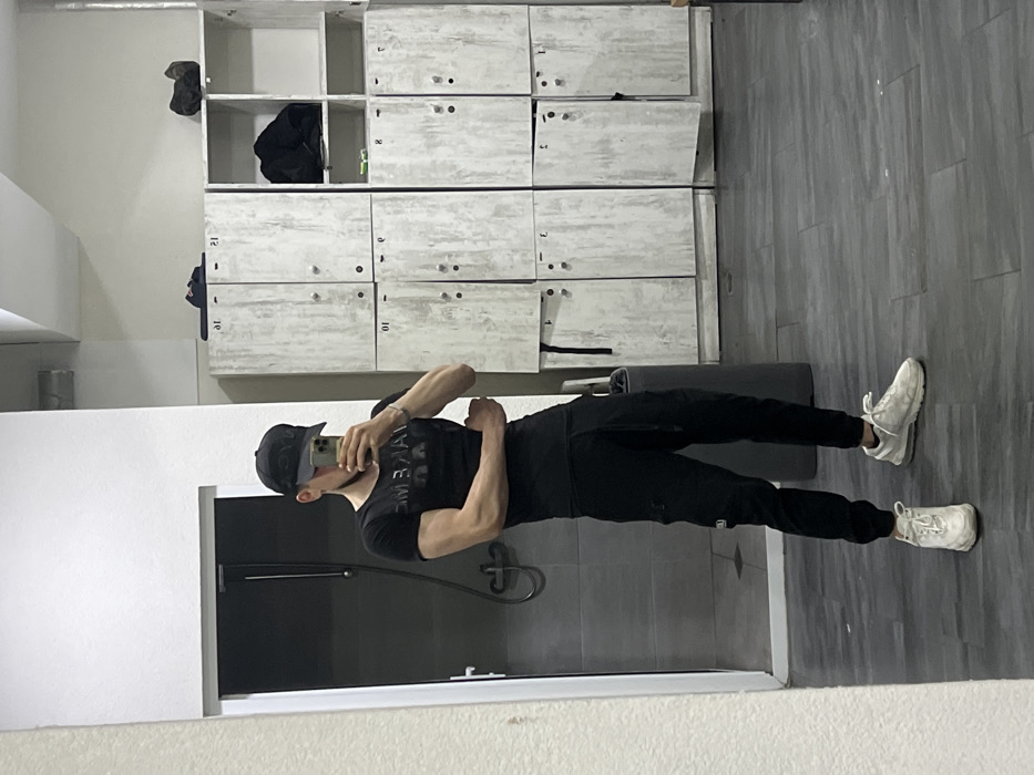 An adult male stands in what appears to be a gym locker room or changing area in Kardzhali, Bulgaria. The subject is capturing a mirror selfie with a smartphone held in his right hand, angled to photograph his reflection. He is dressed in a black baseball cap worn backward, a black short-sleeved t-shirt with a faint dark graphic, black athletic joggers, and white athletic shoes. His left arm is bent at the elbow, with the forearm held horizontally across his midsection.
The environment features a white wall to the left with an open doorway leading to a dark shower stall, where a chrome shower fixture is visible. To the right of the subject, a wall-length installation of light-colored, distressed laminate lockers is present. These lockers are compartmentalized, with several doors bearing numbers (e.g., "10", "01", "21") and some left ajar, revealing dark interiors. Items such as dark sandals and clothing are visible on top of and inside some locker compartments. A dark cylindrical waste receptacle is partially visible between the subject and the lockers. The floor is covered with dark grey rectangular tiles, and the area is illuminated by overhead lighting.
