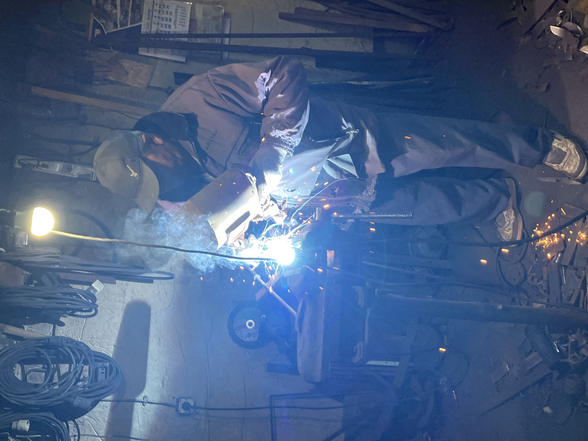 The image depicts a person actively engaged in welding within a dimly lit workshop or industrial setting. The central subject, wearing a cap, a dark jacket, and trousers, is bent over a workpiece, operating a welding gun. A bright, intense arc of light emanates from the welding point, accompanied by numerous hot sparks flying outwards and a plume of smoke rising from the work area.
The environment appears cluttered, characteristic of a working space. On the left, a bare light bulb illuminates a section of the wall where several coiled electrical cables are stacked. Various metal scraps, tools, and equipment, including what looks like a grinding wheel or cutting disk, are visible in the background and foreground, contributing to the industrious atmosphere.
In the upper background, a calendar is visible on the wall, displaying numbers and what appear to be Chinese characters. The overall lighting suggests an indoor scene, potentially during the evening or night, with the welding arc and the single bulb serving as the primary light sources. The person's face is mostly obscured by the cap and the brightness of the welding, so no emotions are discernible. The image captures a moment of intense manual labor and metal fabrication.