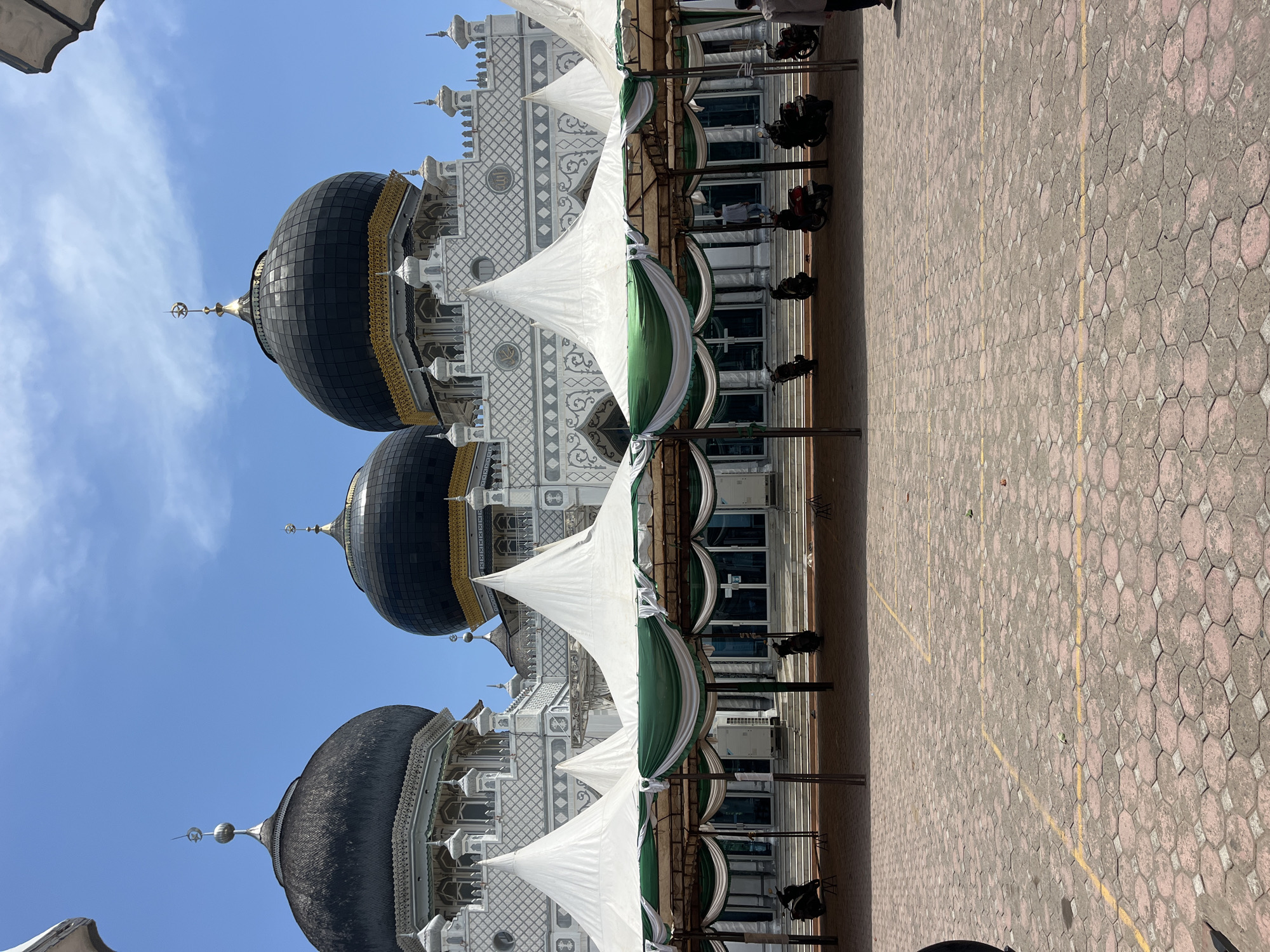 The image depicts the Baiturrahman Keudekruenggeukueh Mosque in Keudekruenggeukueh, Indonesia. The mosque has three visible black domes with gold trim. The structure is predominantly white with geometric patterns. White and green awnings are positioned adjacent to the building. A brick-paved ground is in the foreground. The sky is blue with scattered clouds. The photo is taken at an oblique angle.