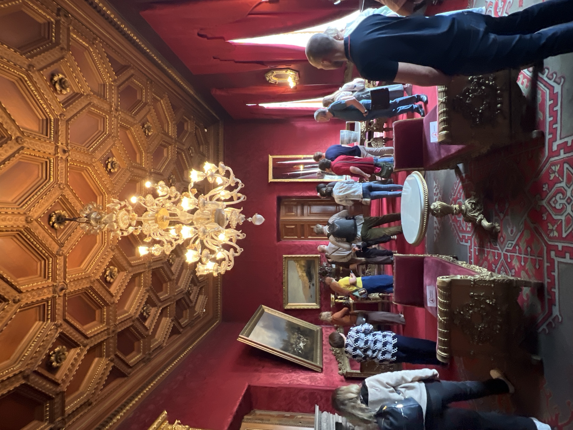 The image depicts a group of tourists viewing an ornate room, likely within a palace or museum, in Saint Petersburg, Russia. The room is richly decorated with red velvet walls, an elaborate gilded ceiling, and a grand chandelier. Several paintings are displayed on the walls, and antique furniture, including gilded chaise lounges, is present. The tourists, dressed in casual attire, are observing the surroundings and each other. The overall atmosphere suggests a guided tour or a visit to a historical site.