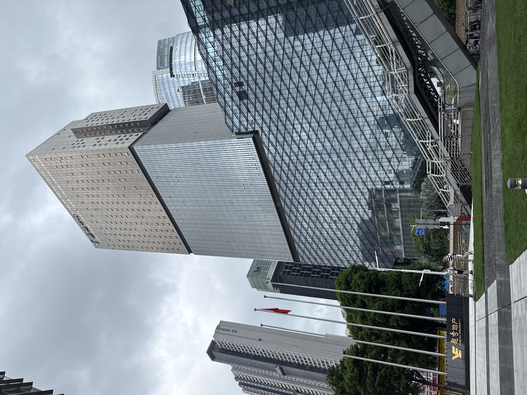 The image shows a cluster of modern skyscrapers in Tianhe District, China. The buildings are characterized by their sleek, glass facades, with one building featuring a distinctive geometric pattern of windows. The sky is overcast, suggesting a cloudy day. In the foreground, a grassy area and a paved walkway are visible, along with a sign that reads "Yuexiu Property" in both Chinese and English. Several flagpoles stand near the sign, with one flag appearing red. A few people are casually walking by, seemingly unaware of the photographer. The overall scene depicts a typical urban environment with contemporary architecture and clear signage indicating a commercial area. The perspective is from a low angle, looking up at the imposing structures.