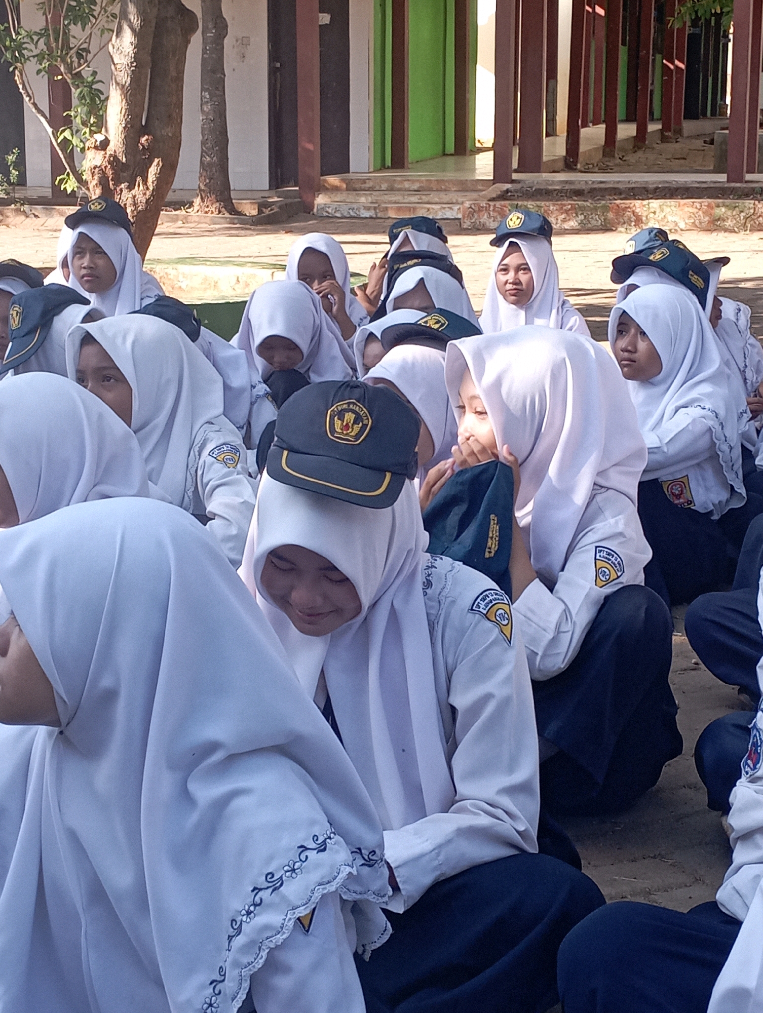 In Banyuurip, Indonesia, a group of students wearing white hijabs and uniforms sit on the ground in front of a building with maroon pillars and green walls. Most of the students are looking down, some with their hands to their faces. One student in the foreground is looking up and to the side. Several students are wearing dark blue caps with gold emblems. The ground is dirt and gravel.
