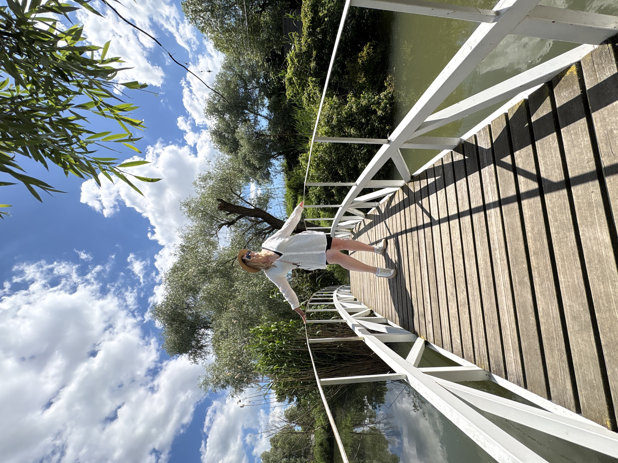 An outdoor scene is depicted from an unusual, rotated perspective, showing a woman on a wooden bridge under a bright sky. The woman, with blonde hair, is wearing a hat, sunglasses, a white long-sleeved shirt, black shorts, and white sneakers. She stands on the bridge with her arms outstretched, leaning back, appearing to balance or pose for the camera.
The bridge features dark wooden planks for the walkway and a white, angular railing on one side, extending diagonally across the frame. Shadows from the railing and the woman are sharply cast on the bridge planks, indicating direct sunlight and a clear day. To the right of the bridge, a body of water (likely a river or canal) is visible, reflecting parts of the sky and surrounding greenery. On the left side, lush green trees and dense foliage line the water's edge, creating a natural backdrop. Above and below the bridge, a blue sky dotted with scattered white clouds fills the background. No text is visible. The scene suggests a relaxed moment in a scenic natural environment, specifically Gay-Kodzor, Russia.