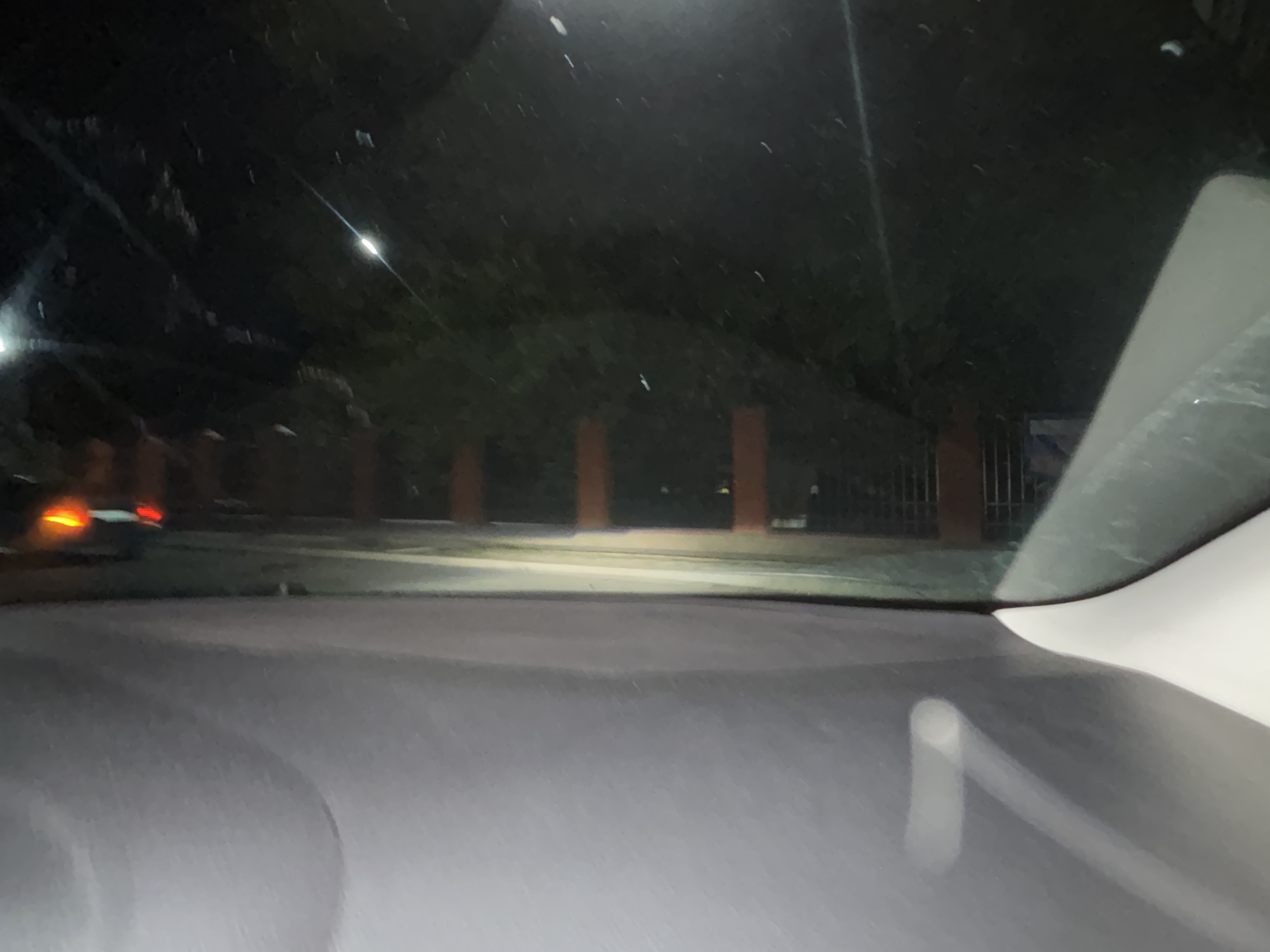 The image, taken at night from inside a vehicle, captures a dimly lit outdoor scene. The perspective suggests the viewpoint is from the driver's seat looking forward through the windshield. The foreground is dominated by the dark, reflective surface of the car's dashboard.
In the middle ground, the headlights of the vehicle illuminate a paved surface that appears to be a road or pathway. Beyond this, a dark wall or fence is visible, with vertical orange posts spaced intermittently. Trees are sparsely visible behind the fence, their foliage appearing dark and indistinct in the low light.
Further in the distance, on the left side of the frame, the taillights of another vehicle are visible, emitting a bright red glow. These lights are the most distinct artificial illumination besides the car's headlights.
The overall impression is one of a quiet, possibly residential or suburban, street at night. There are no people clearly visible in the frame. The environment appears to be an urban setting in Grozny, Russia, based on the provided context. The scene lacks any specific architectural details that would definitively pinpoint a more precise location within the city. The image's quality, with its low light conditions, leads to a somewhat blurry and indistinct depiction of the surroundings.
