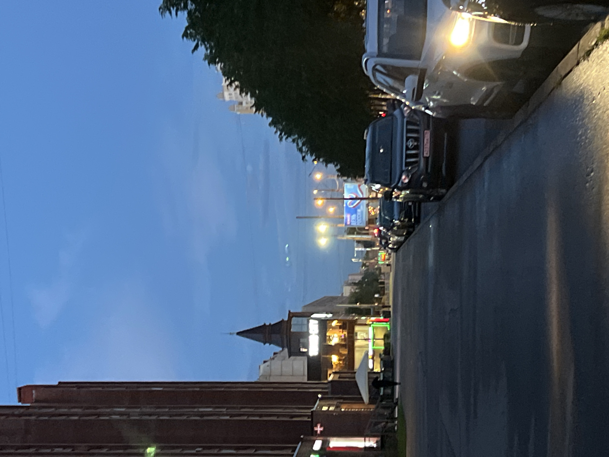The image depicts a street scene in Saint Petersburg, Russia, during twilight. The sky is a deep blue, suggesting the end of the day. On the right side of the frame, cars are parked along the curb, their headlights illuminating the wet asphalt. One car, a white SUV, has a bright yellow headlight prominently visible. Another dark SUV, possibly a Mercedes-Benz based on the grille design, is parked behind it, displaying a license plate with Cyrillic characters.
Further down the street, buildings line the left side. A brick building occupies the lower left portion of the image. To its right, a building with a distinctive pointed, dark brown roof structure stands out. This building appears to be a commercial establishment, with illuminated windows showing warm, yellow light. Signs with colorful neon lights, including a red cross symbol on a white background, are visible, indicating shops or services.
The overall atmosphere is that of a quiet evening in an urban environment. The wet road surface suggests recent rain, which is common in Saint Petersburg. The lighting is a mix of natural twilight and artificial lights from the street and buildings, creating a somewhat moody and atmospheric scene. No specific activities or events are clearly taking place, but the presence of parked cars and illuminated buildings suggests normal evening activity. The text "ДЕНЬ РОССИИ" (DEN' ROSSII), meaning "Russia Day," is visible on a blue sign in the distance, indicating that the photograph might have been taken around June 12th.