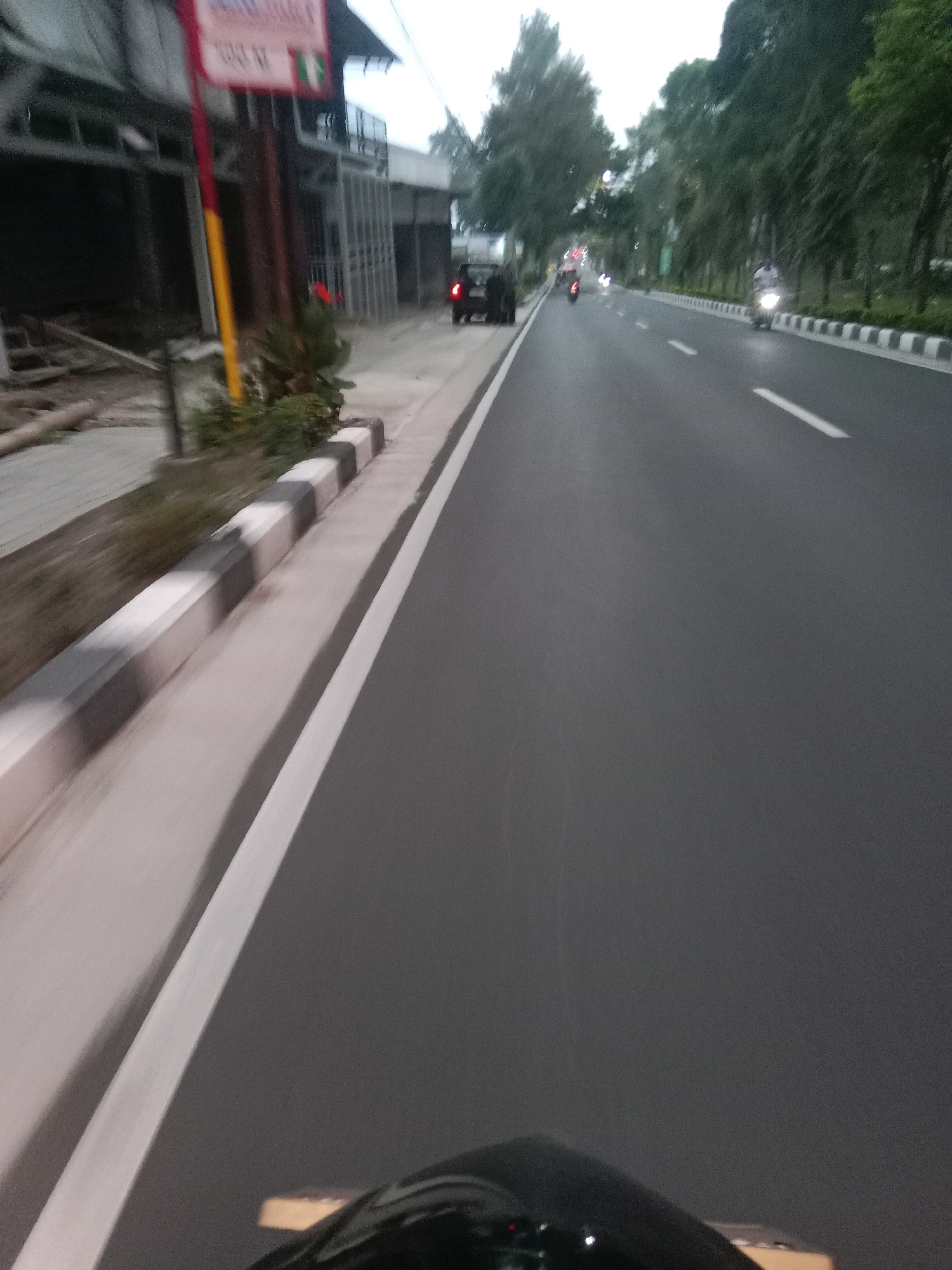 The image shows a street scene in Kalianget, Indonesia. The perspective is from the viewpoint of someone riding a motorcycle, with the front of the motorcycle visible in the foreground. The road is paved with asphalt and has white lane markings. On the left side of the road, there is a sidewalk with a curb that has alternating black and white stripes. Beyond the sidewalk, there are buildings under construction, with exposed metal framing and some debris. A red and yellow pole stands near the buildings. Further down the road, several vehicles are visible, including a black SUV and motorcycles. Trees line the right side of the road, and the sky appears overcast. The time of day appears to be dusk or early morning.