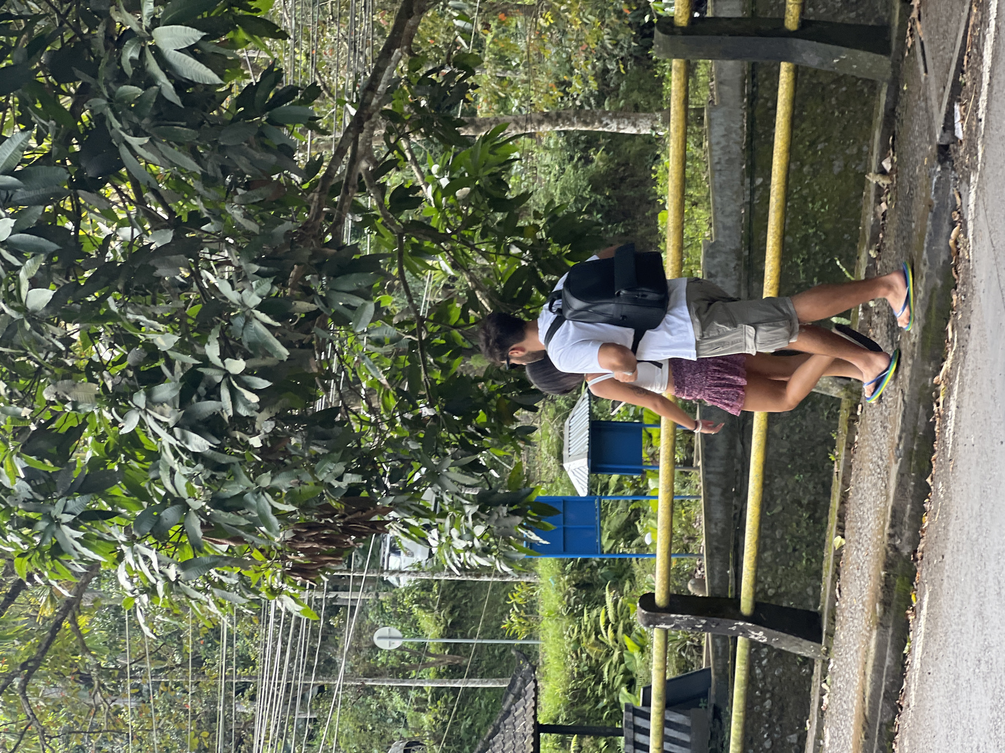 In Tampaksiring, Indonesia, two individuals, a man and a woman, walk side-by-side along a paved path beside a yellow railing. The man, in a white t-shirt and grey cargo shorts, wears a black backpack. The woman, in a white tank top and a short, patterned skirt, walks to his left. They are both wearing flip-flops. The path is bordered by a concrete barrier and a yellow metal railing. Behind the railing, lush green vegetation and trees dominate the landscape, with some power lines visible in the background. A small thatched-roof structure and a blue information booth are situated further back. The scene appears to be outdoors, possibly near a natural attraction or a roadside area in a tropical environment.