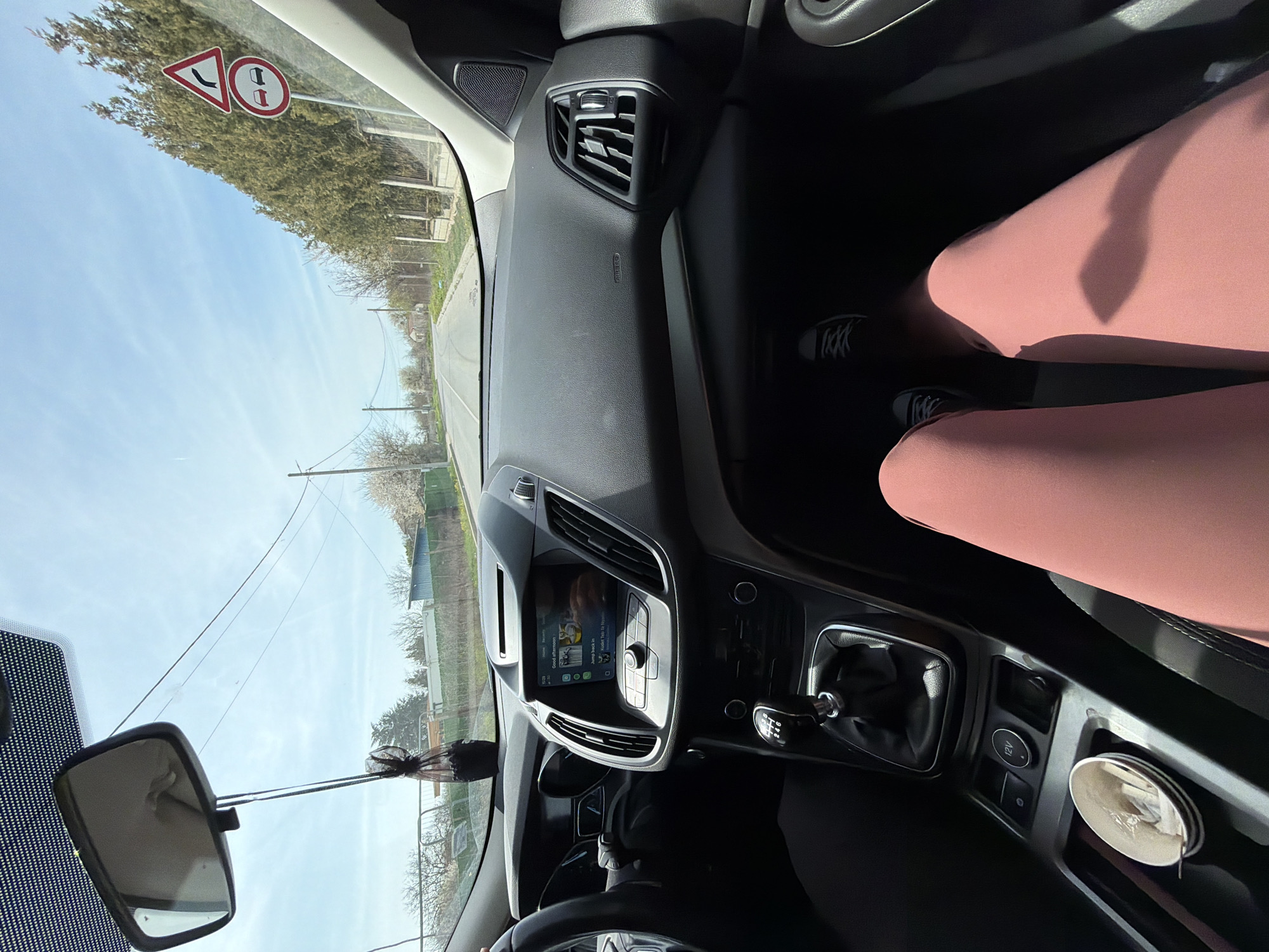 The image, likely taken from inside a vehicle, shows the view through the windshield of a road near Kavarna, Bulgaria. A road sign indicating a sharp left turn ahead is visible on the left. Inside the vehicle, the dashboard, steering wheel, gear shift, and center console are visible. A pair of legs clad in rose-colored leggings and black sneakers are also visible on the passenger side. A rearview mirror hangs from the windshield. A cup sits on the center console. The sky is clear.