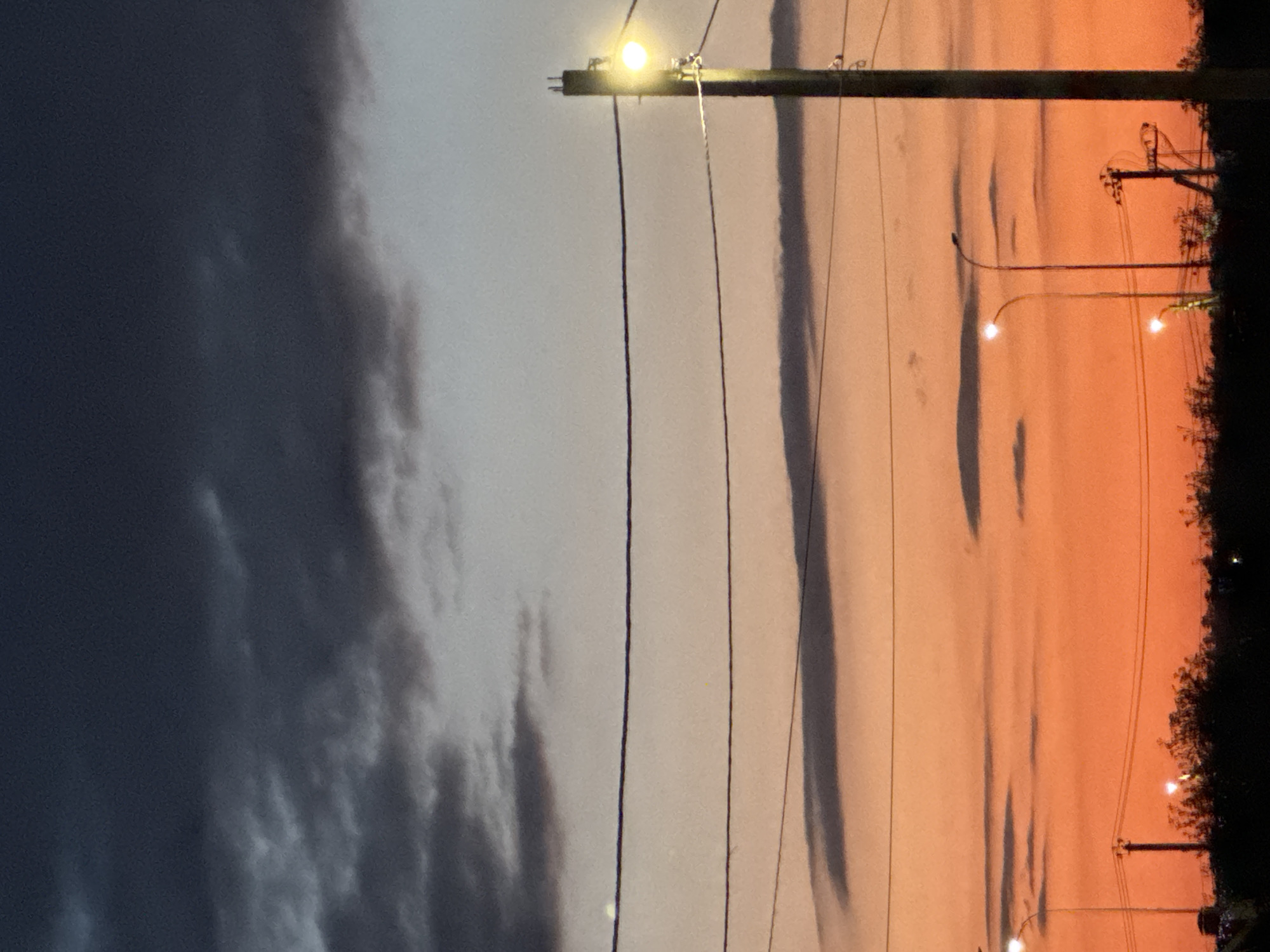 This is a vertical, low-light photograph of a dramatic sunset. The scene is divided into two distinct halves by a dark, overcast sky on the left and a vibrant orange and pink sky on the right. Multiple power lines stretch across the frame, originating from a utility pole on the right side of the image. The light from the setting sun casts a warm glow on the right side, illuminating silhouetted trees and appearing to reflect on a surface, possibly water, below. There are a few streetlights visible, some of which are illuminated. The left side of the image is dominated by dark, moody clouds, suggesting impending or ongoing weather. The overall mood is one of transition, with the stark contrast between the light and dark skies creating a sense of awe and perhaps foreboding. The time of day is clearly dusk. No people or text are visible in the image.