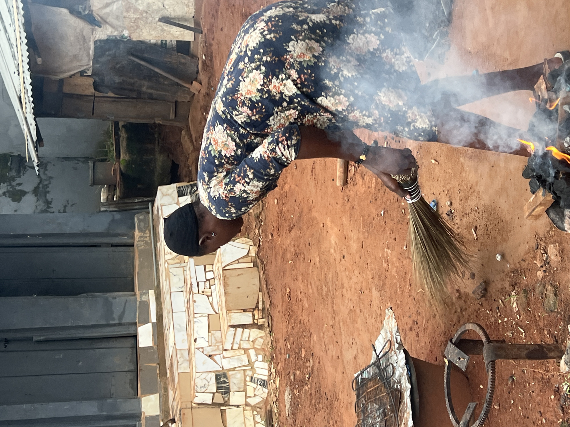 In Nkpor, Nigeria, a woman wearing a dark floral print dress and a black head covering sweeps with a broom near a small fire. The fire burns on the ground, surrounded by charcoal and wood. The ground is made of reddish-brown dirt. In the background, there is a building with a corrugated metal roof. To the left of the woman, there is a metal grill situated on top of a large, dark cylindrical object, possibly a drum. A decorative tiled structure is visible behind the grill. The scene appears to be outdoors, in an area with elements of both domestic activity and the surrounding environment.