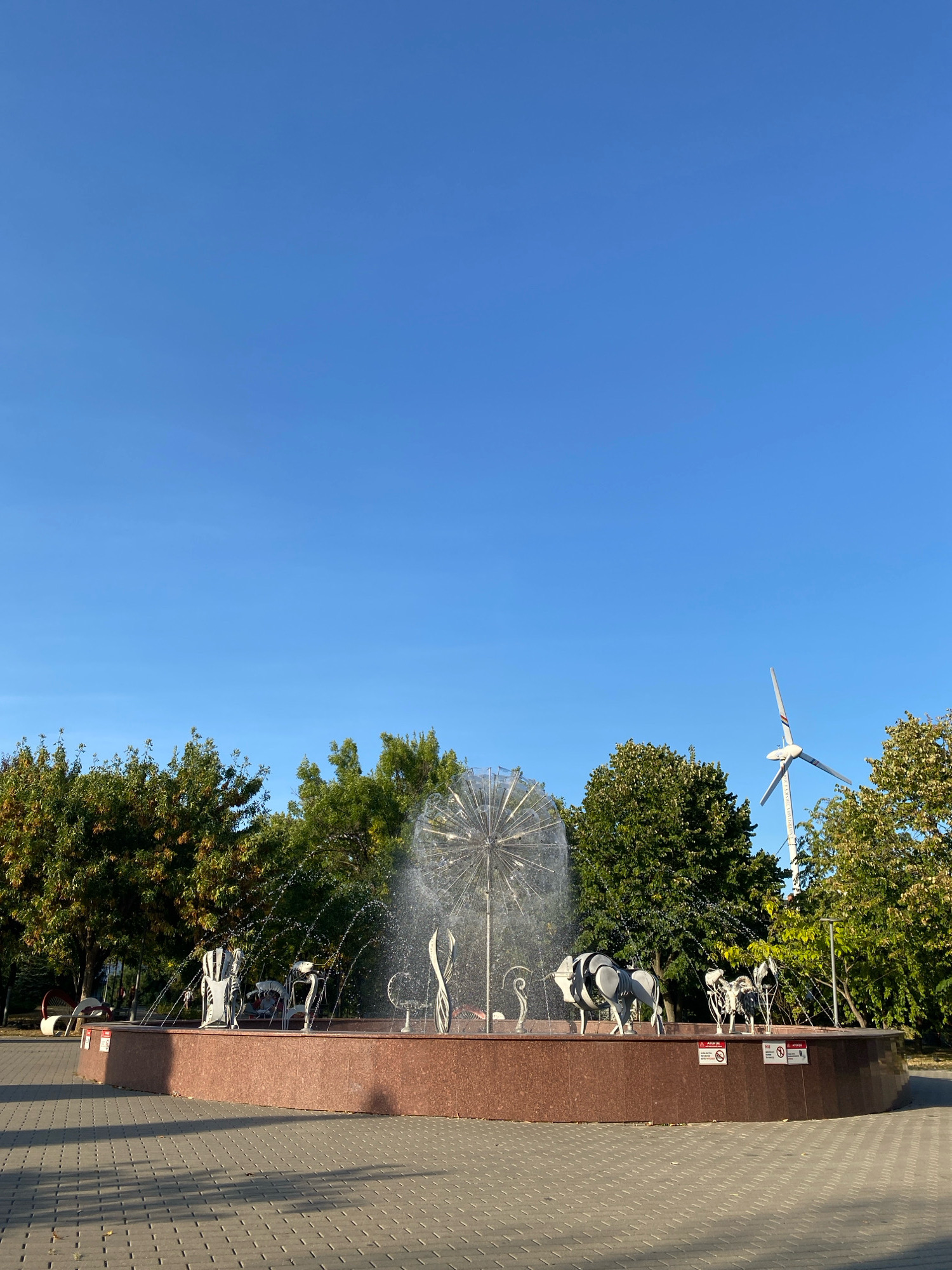 The image shows a fountain with metallic sculptures of animals in a park in Chișinău, Moldova. The fountain is decorated with a water display resembling a dandelion. In the background, there are trees and a wind turbine. The ground is paved with grey tiles, and the sky is clear and blue. There are no people visible in the image.