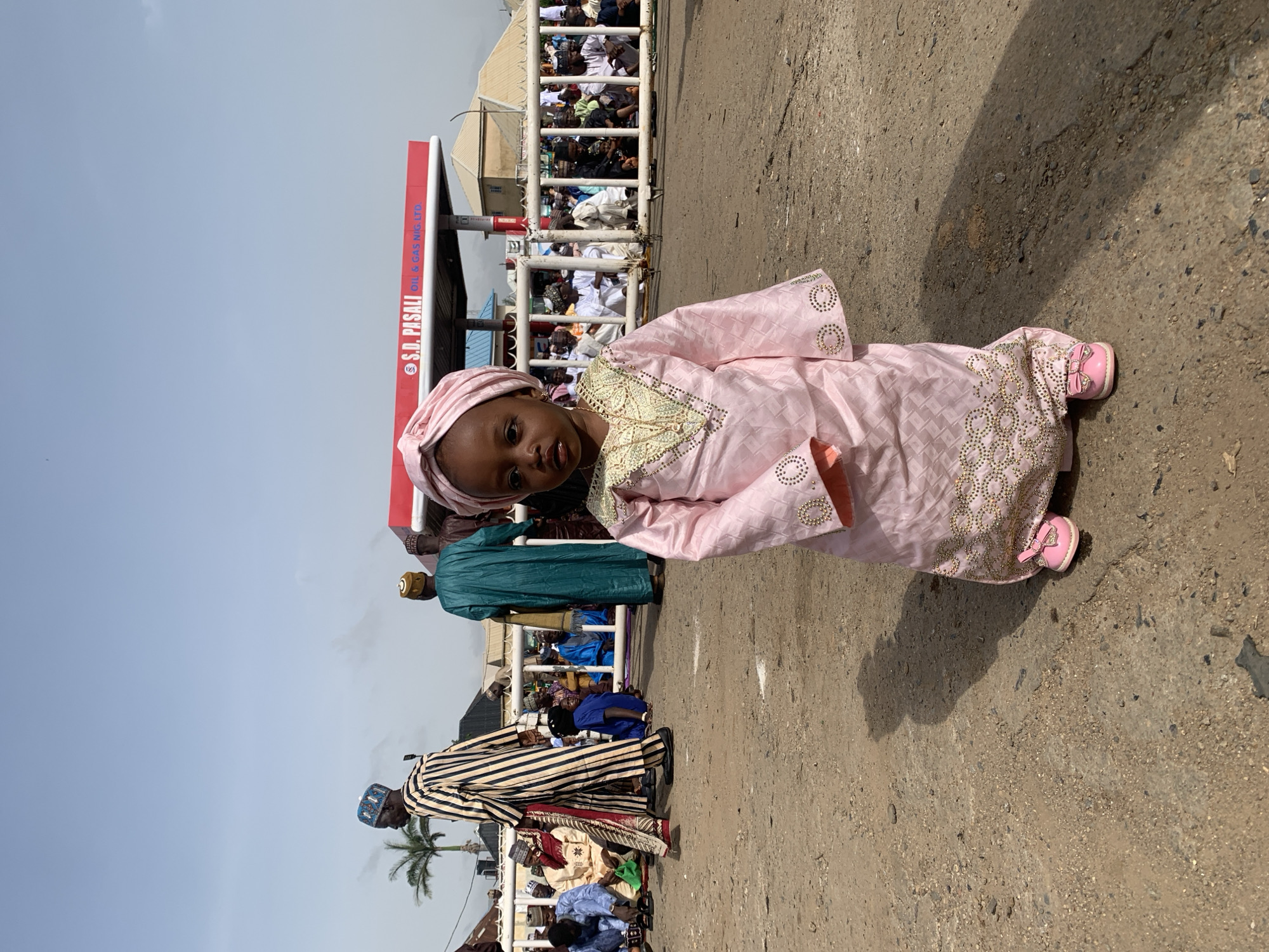 A young child wearing a pink traditional outfit stands in front of a crowd of people behind a white metal barrier. A red sign with white lettering is visible on the left side of the frame. The child is wearing a matching pink head wrap and pink shoes. The ground is concrete. The photograph may have been taken in Jos, Nigeria.