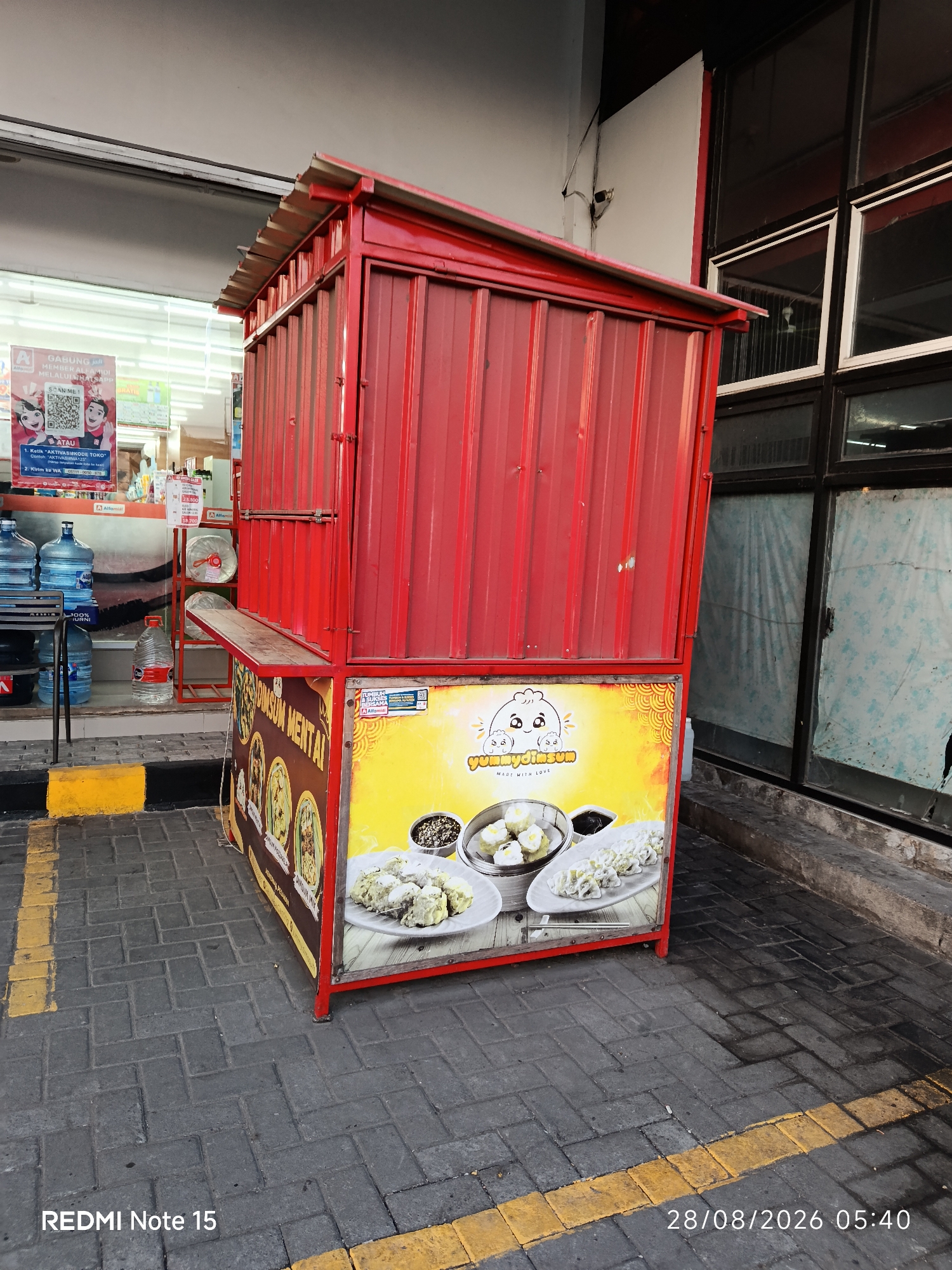 A red food cart is parked on a grey brick sidewalk in front of a building with large windows. The food cart has a corrugated metal roof and sides, and a wooden serving counter that is slightly ajar. A colorful sign on the front of the cart displays images of dim sum and the text "yummydimsum MADE WITH LOVE". The building behind the cart appears to be a store or commercial establishment, with shelves visible through the windows. Several large water jugs are stacked to the left of the food cart. Yellow lines are painted on the sidewalk. The scene appears to be outdoors, possibly in a public area of Keboansikep, Indonesia, given the watermark "REDMI Note 15" and the date "28/08/2026 05:40". There are no people visible in the image.
