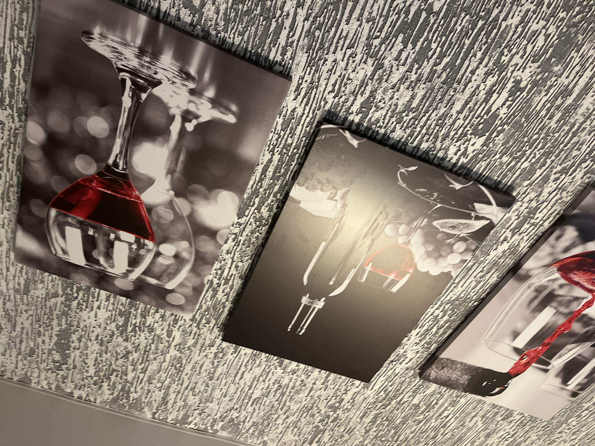 The image displays three rectangular art prints mounted on a heavily textured, light gray and white wall. The perspective is angled upwards, showing part of a ceiling or upper wall edge at the bottom. This is an indoor setting, potentially in Chaikovsky, Russia, given the provided context.
All three prints feature wine-related subjects, rendered primarily in grayscale with the wine itself highlighted in a vibrant red color.
The leftmost print shows an inverted wine glass with red wine, set against a blurred, bokeh-lit background. The glass is shiny, reflecting light.
The central print presents two inverted wine glasses, one larger and one smaller, with red wine visible in the smaller glass. A cluster of grapes is faintly visible in the background against a dark canvas.
The rightmost print captures a dynamic scene of a stream of red wine being poured into an inverted wine glass. A dark wine cork or corkscrew element is visible near the bottom left of this print.
The wall's texture is rough and layered, creating a relief pattern. No people are visible, nor is any specific activity taking place other than the static display of the artwork. There is no visible text in the image. The overall impression is one of decorative interior design, likely in a home or a commercial establishment with a wine theme.