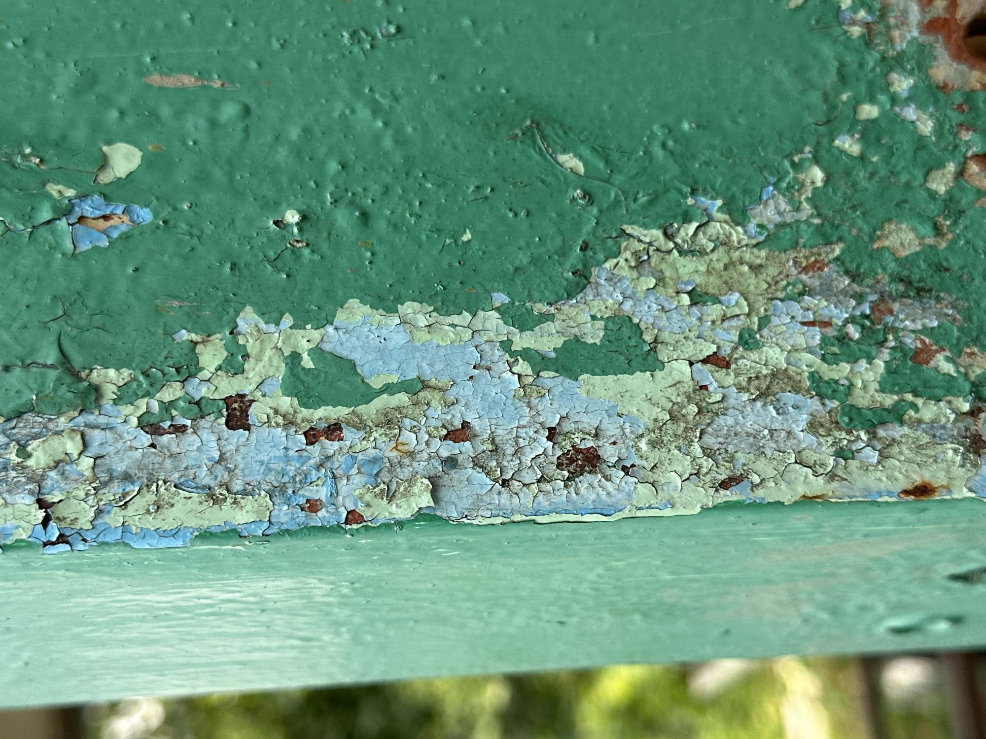 The image is a close-up, high-angle shot of a weathered green metal surface, likely a railing or structural element. The surface exhibits significant paint peeling and rust. Multiple layers of paint are visible, including shades of green, light blue, and a reddish-brown rust color. The paint is cracked and flaking, revealing the underlying metal and corrosion. The texture is rough and uneven due to the peeling paint and pitting of the metal. The background is softly blurred, showing out-of-focus green foliage and hints of yellow, suggesting an outdoor environment. There are no people or discernible actions depicted. The scene is static and focuses on the textural and color details of the decaying paint. The specific location in Yessentuki, Russia cannot be determined from the image.