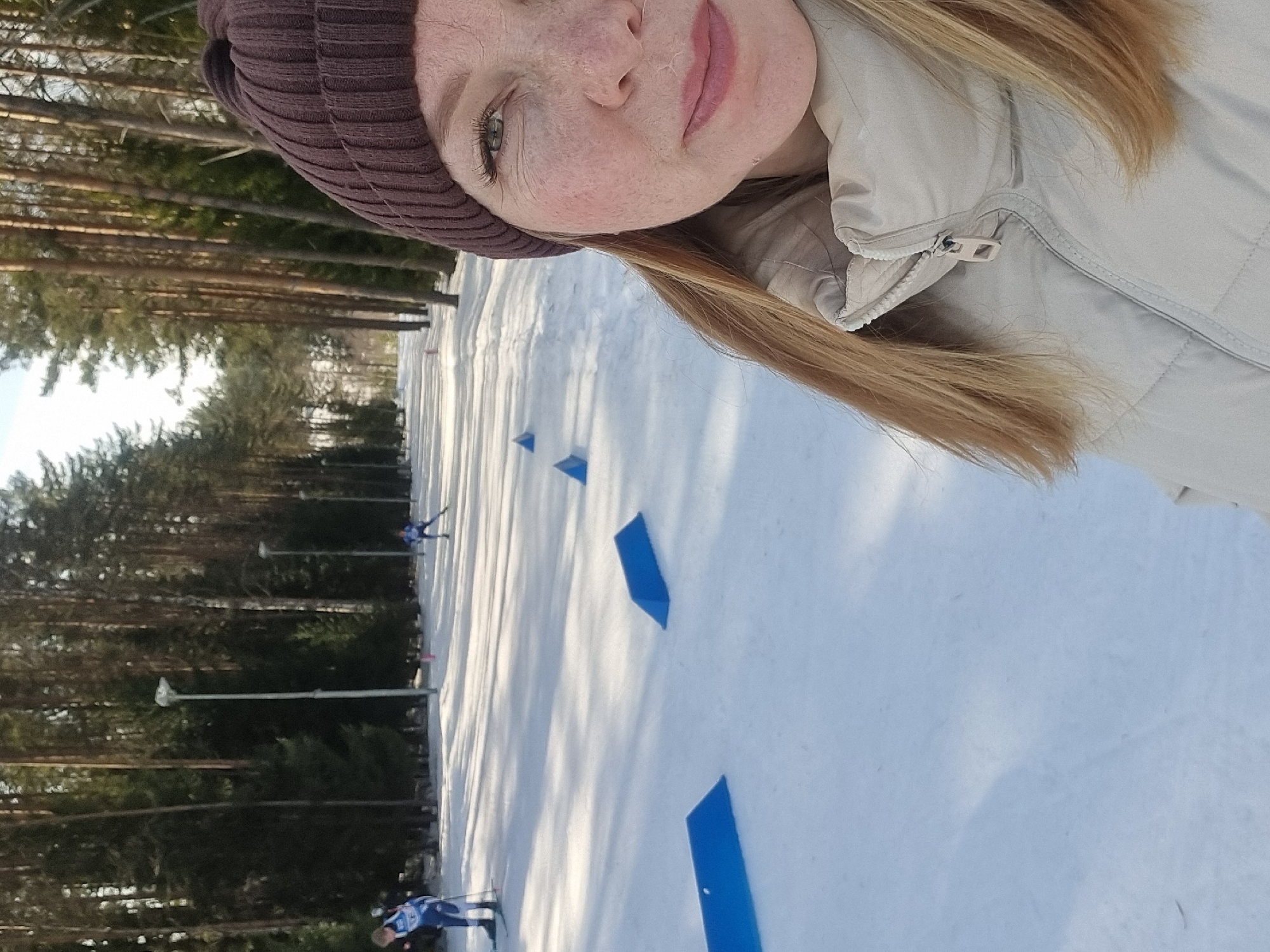 The image depicts an outdoor winter scene on a snow-covered ski track in Прикамский, Russia. The foreground, positioned on the right side of the frame, shows the right half of a female subject's face and upper body. She has fair skin with visible freckles, light-colored eyes, and light brown hair partially covered by a dark brown ribbed knit beanie. She is wearing a light beige or cream-colored padded jacket with a partially zipped collar.
The background features a wide, snow-covered path, indicative of a ski track, extending into the distance. This path is bordered on both sides by tall pine trees. Several blue, triangular markers are visible on the snow, arranged along the track. In the midground, two individuals are present on the track. One person on the far left is wearing a blue and white ski suit and holding ski poles, appearing to be standing. A second person, further down the track on the right, is actively cross-country skiing, also dressed in a blue ski suit. Tall streetlights are intermittently visible along the treeline. Sunlight casts distinct shadows across the snow.