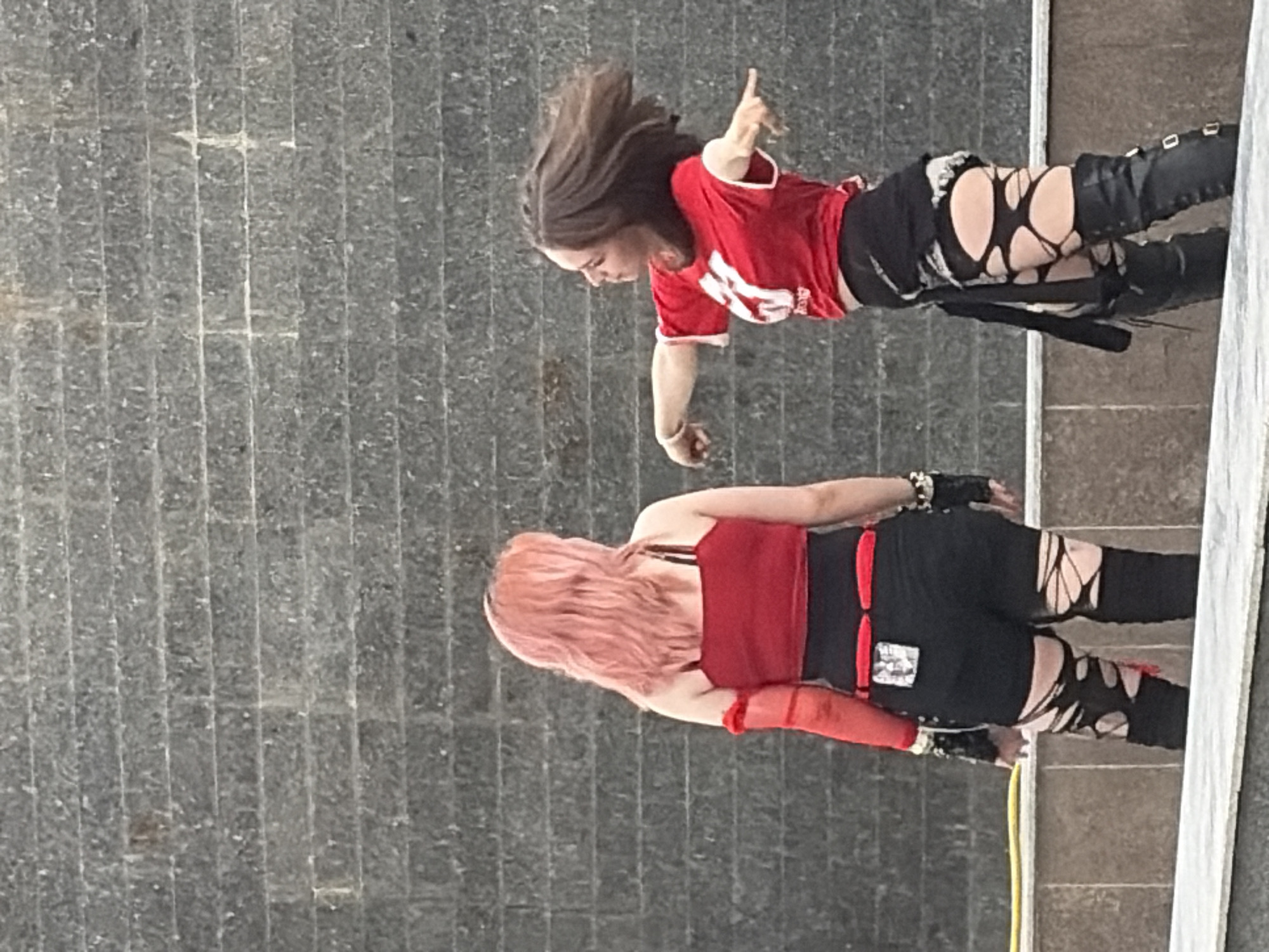 Two young women are standing in front of a dark grey stone wall, possibly a water feature. The woman on the left has long, wavy pink hair and is wearing a red tank top, black shorts with torn details, and black thigh-high stockings with similar tear details. She also has red arm coverings. The woman on the right has dark brown hair and is wearing a red t-shirt with white lettering, black shorts, and black thigh-high boots with buckle details. Her hair is blurred, suggesting movement, and her arms are outstretched as if dancing or posing. A yellow hose lies on the ground in front of them.
