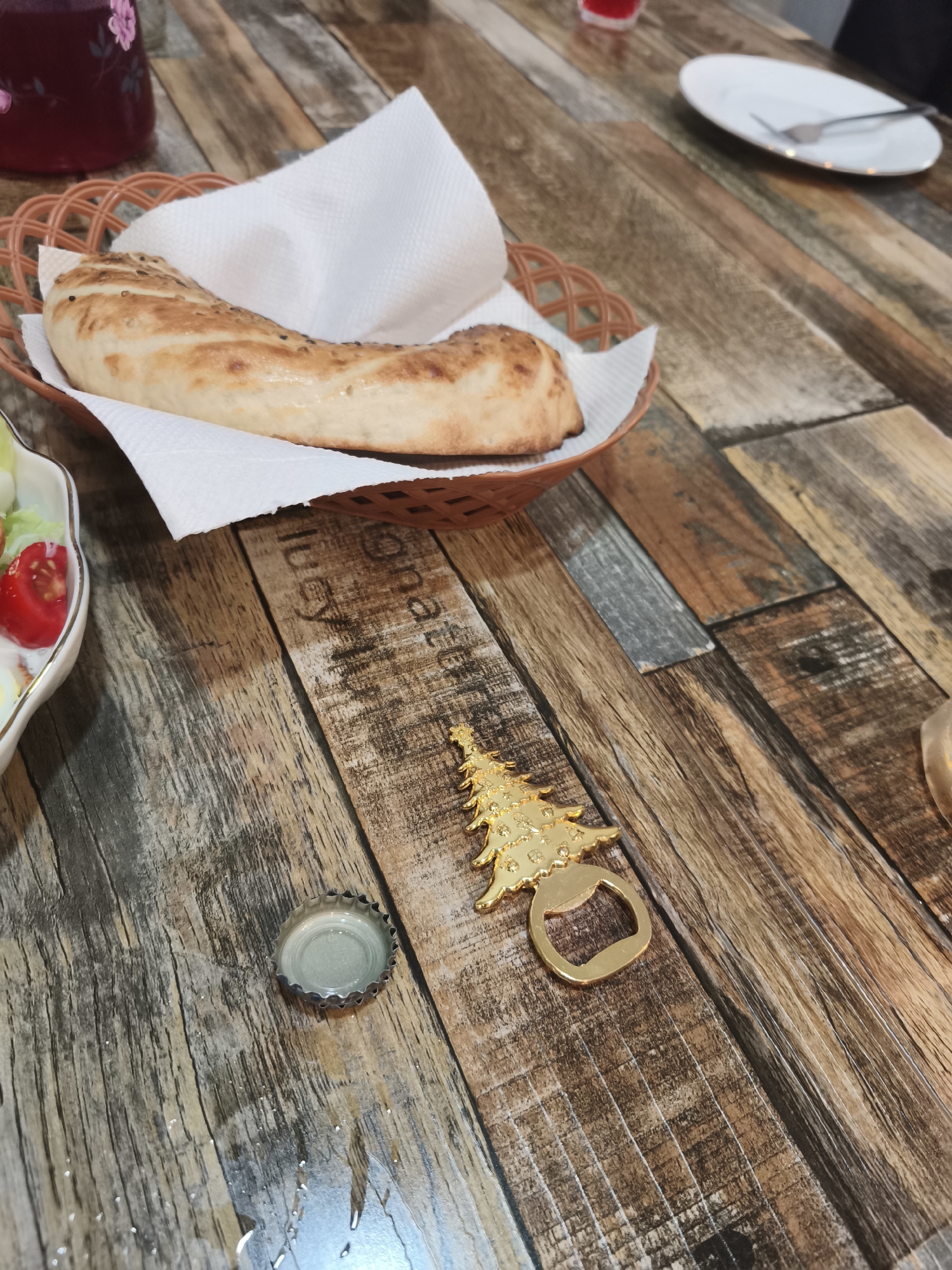 A close-up, eye-level shot captures a wooden table with food and a bottle opener. In the upper left corner, a dark red beverage is visible in a glass container, with a floral pattern on its surface. Next to it, a brown woven basket holds a piece of bread, possibly lavash or flatbread, topped with sesame seeds and resting on a white napkin. To the left of the basket, a white dish contains a salad with cherry tomatoes, lettuce, and what appears to be boiled eggs and dressing.
Dominating the center of the frame, a bottle opener shaped like a golden Christmas tree is positioned on the wooden table. To its left, a silver bottle cap lies on the surface, near a small puddle of liquid. The wooden table itself exhibits a rustic, textured surface composed of varying shades of brown and gray planks. Faint text, possibly a brand logo or decoration, is visible etched into the wood grain. The background is blurred, indicating a shallow depth of field, with a hint of a white plate and fork on the right side of the image. The scene appears to be set indoors, likely in a restaurant or dining area, given the presence of food and tableware. The location is unspecified as Asan-si, South Korea, though this information cannot be confirmed from the visual elements alone.