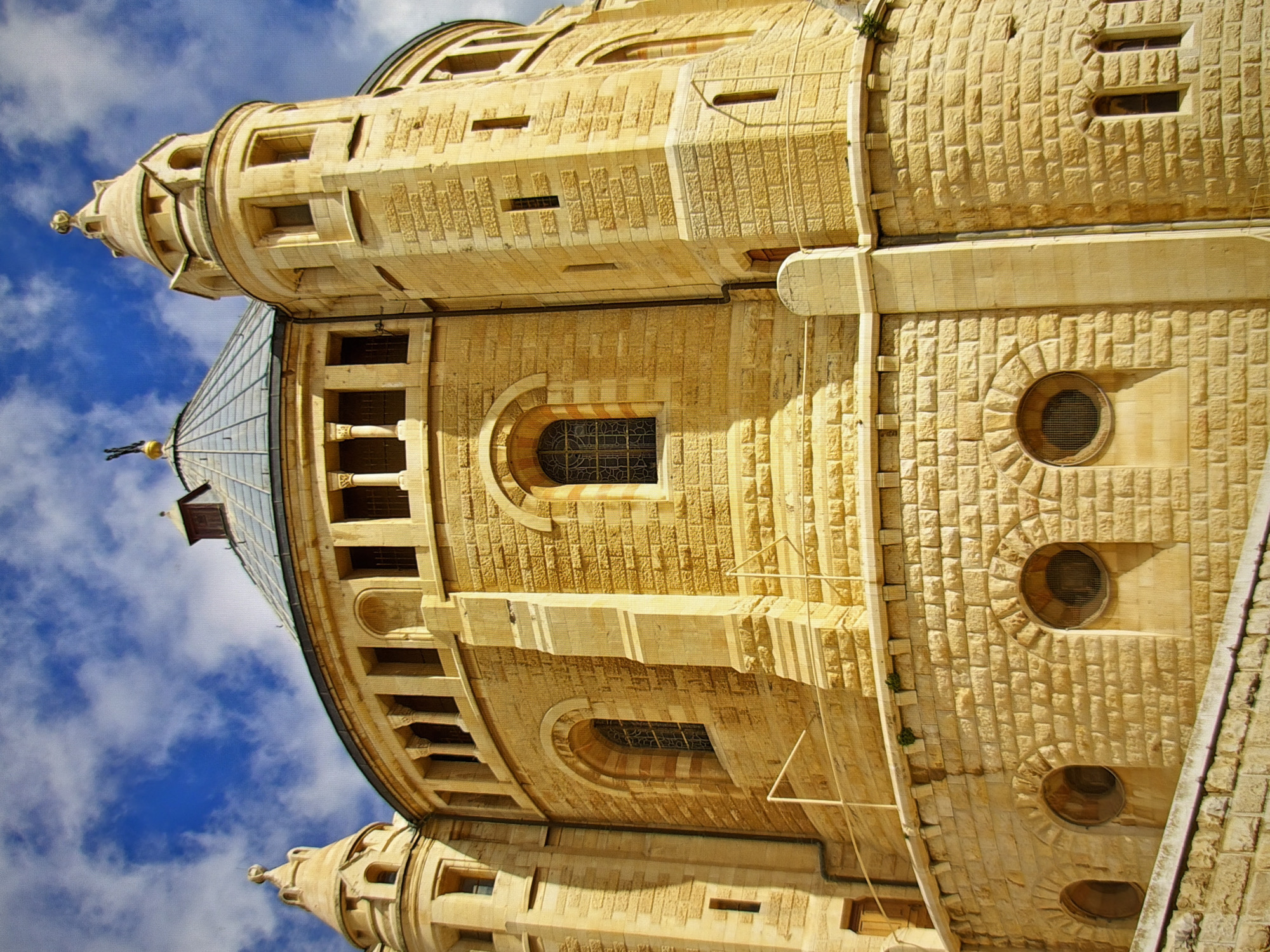 This is a low-angle, rotated-right shot of a stone building with a blue sky and clouds in the background. The building appears to be made of light-colored, roughly cut stone blocks, stacked and arranged in patterns. It features multiple architectural elements, including a tall, conical spire topped with a decorative finial, a rounded, glass-paneled roof section, and various windows and circular openings. The building's facade is detailed with carved patterns and recessed areas, suggesting a historical or religious structure. The lighting indicates it is daytime, with shadows cast by the architectural features. The overall impression is one of grandeur and historical significance. The crops help to focus on the intricate stonework, window details, and the contrast between the building and the sky. Given the provided location context of Kyiv, Ukraine, this building could be a church, monastery, or other significant landmark.