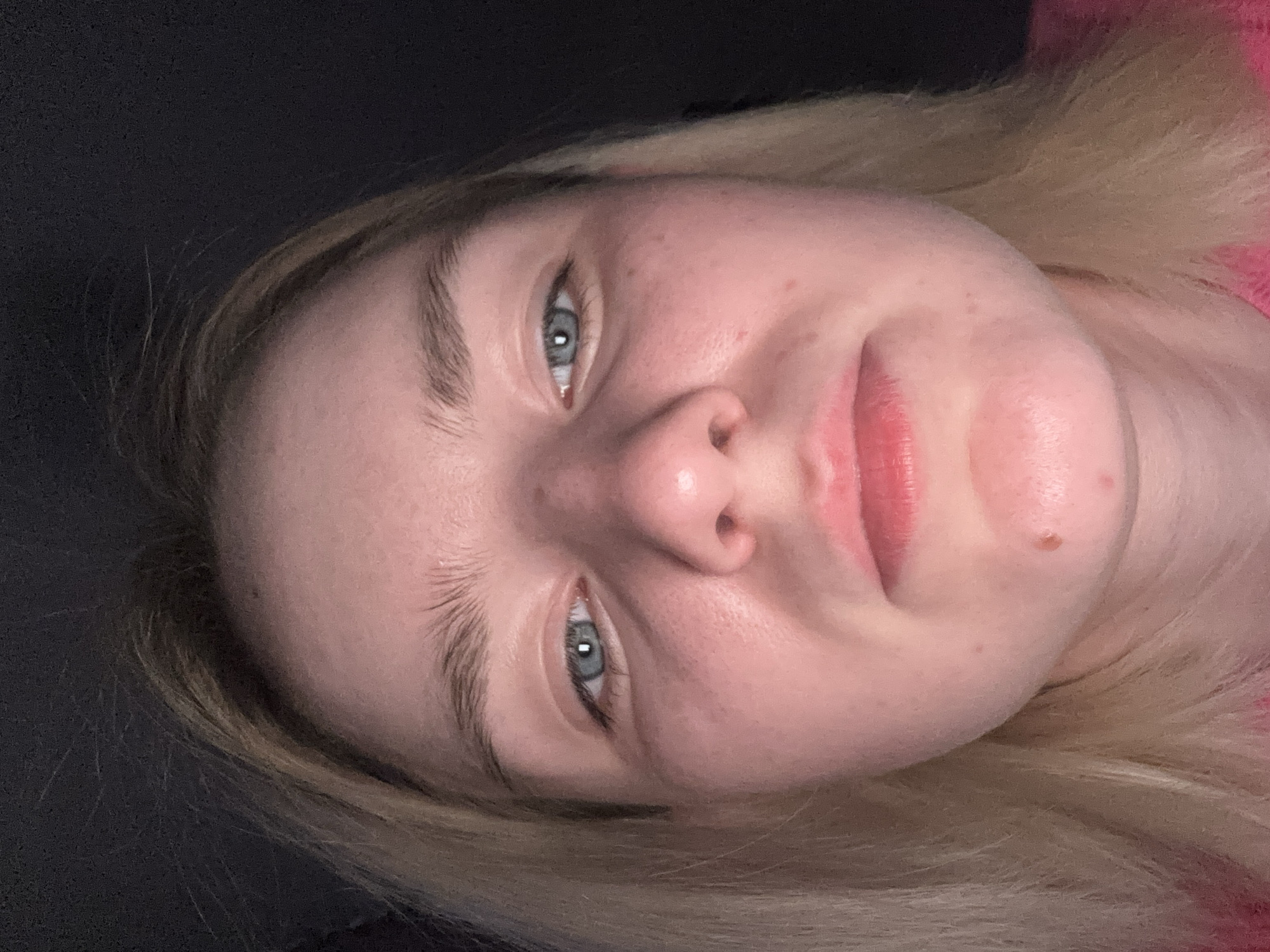 This media file features a close-up portrait of a young woman, likely taken as a selfie, with the image rotated. The primary subject is her face, angled upwards towards the viewer. She has fair skin, light blonde or light brown hair, and striking blue-grey eyes. Her eyebrows are light and natural. Her lips are full, a natural reddish-pink color, and slightly parted. A small mole is visible on her chin, and there are minor skin imperfections like small bumps or redness on her cheeks. Her expression appears neutral, with a slight, subtle smirk.
A hint of pink fabric, possibly from a sweater or top, is visible on the right side of the frame, suggesting her attire. The background is uniformly dark and appears out of focus, indicating an indoor setting or low-light environment. The crops provided confirm these details, showing intricate textures of her hair, the light color of her eyebrows, the details of her blue-grey eyes, and the mole on her chin. No discernible activity or event is taking place beyond posing for the photograph. There is no visible text in the image. The location context provided for this image is Sinyavino, Russia.