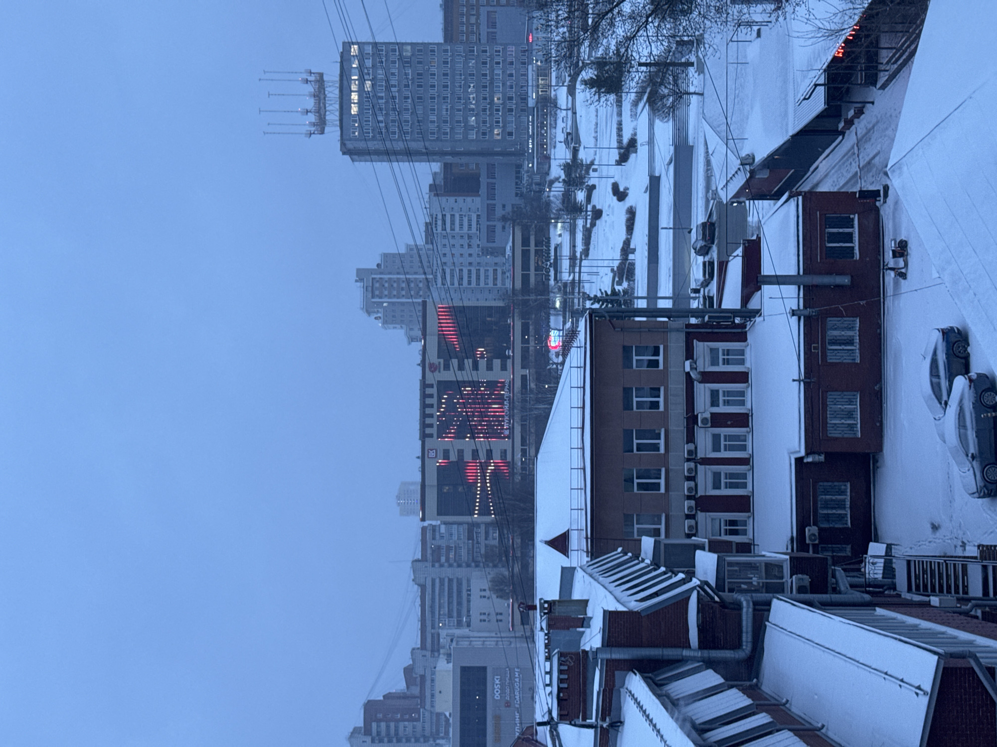 The image shows a cityscape in Perm, Russia, during winter. Snow covers rooftops, streets, and open areas. Tall buildings, including those with lighted displays, form the urban skyline. The sky is overcast. Power lines crisscross the view. A parked car is visible.