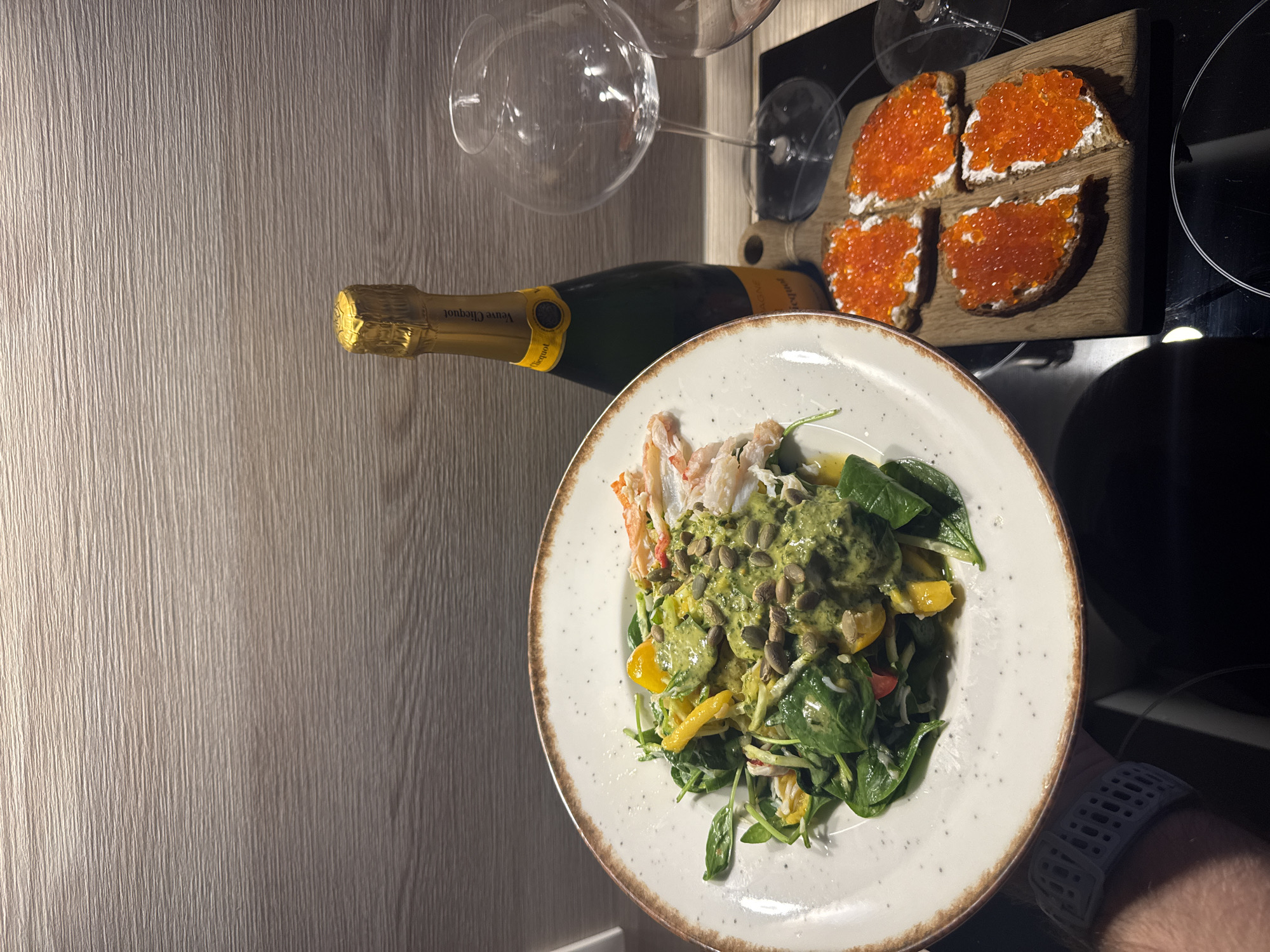 A close-up, high-angle shot shows a meal laid out on a textured, wood-grain countertop in Lednevo, Russia. In the foreground, a white plate with a speckled brown rim holds a vibrant salad composed of dark green spinach, yellow bell peppers, and what appears to be crab meat. The salad is generously drizzled with a green sauce and topped with pumpkin seeds. To the right of the salad, a wooden cutting board displays four small open-faced sandwiches topped with cream cheese and bright red caviar. Behind the food, a green bottle of Veuve Clicquot champagne stands upright, its gold foil cap glinting. Two empty wine glasses are positioned to the right of the champagne bottle. The background features a subtly textured wooden wall. A person's arm wearing a grey smartwatch is visible on the left side of the frame, holding the plate. The overall scene suggests an indoor dining setting, possibly a home or restaurant, with a focus on an elegant meal.