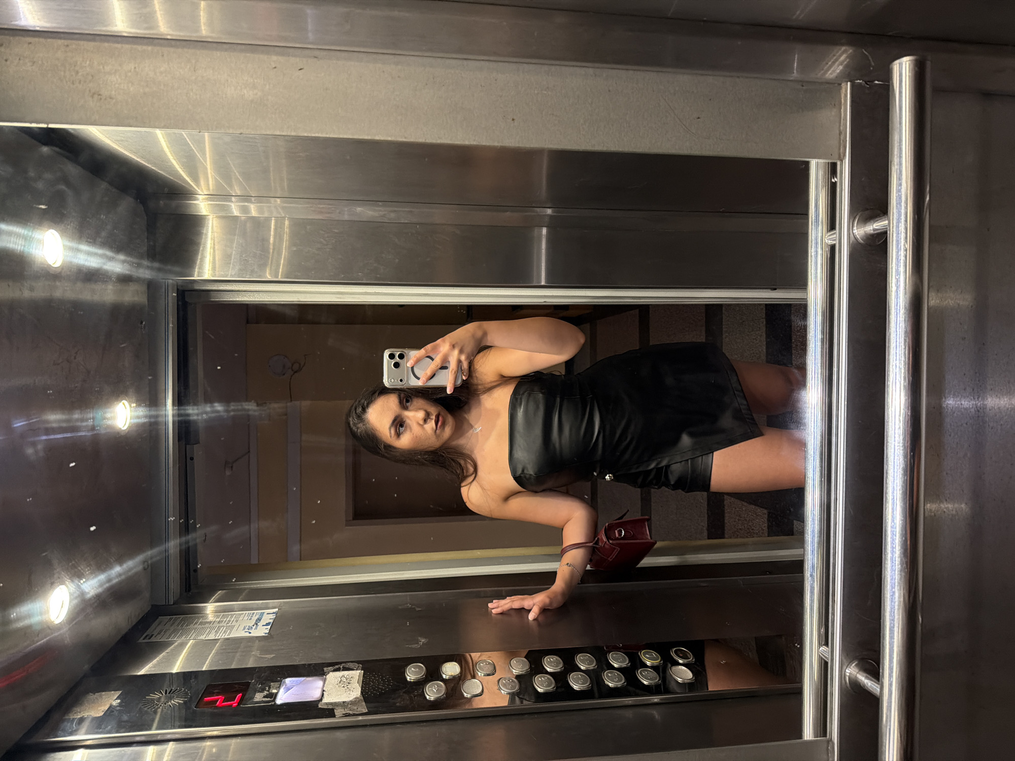 A young woman with long, dark hair stands in an elevator, taking a selfie in the mirror. She is wearing a strapless black leather dress with an asymmetrical hem and a small burgundy handbag. Her left hand rests on the elevator wall, next to a panel of buttons. The elevator walls are made of brushed metal, and the floor appears to be tiled. The lighting is artificial and casts reflections on the metallic surfaces. The image does not contain any information that would indicate the location is Varna, Bulgaria.