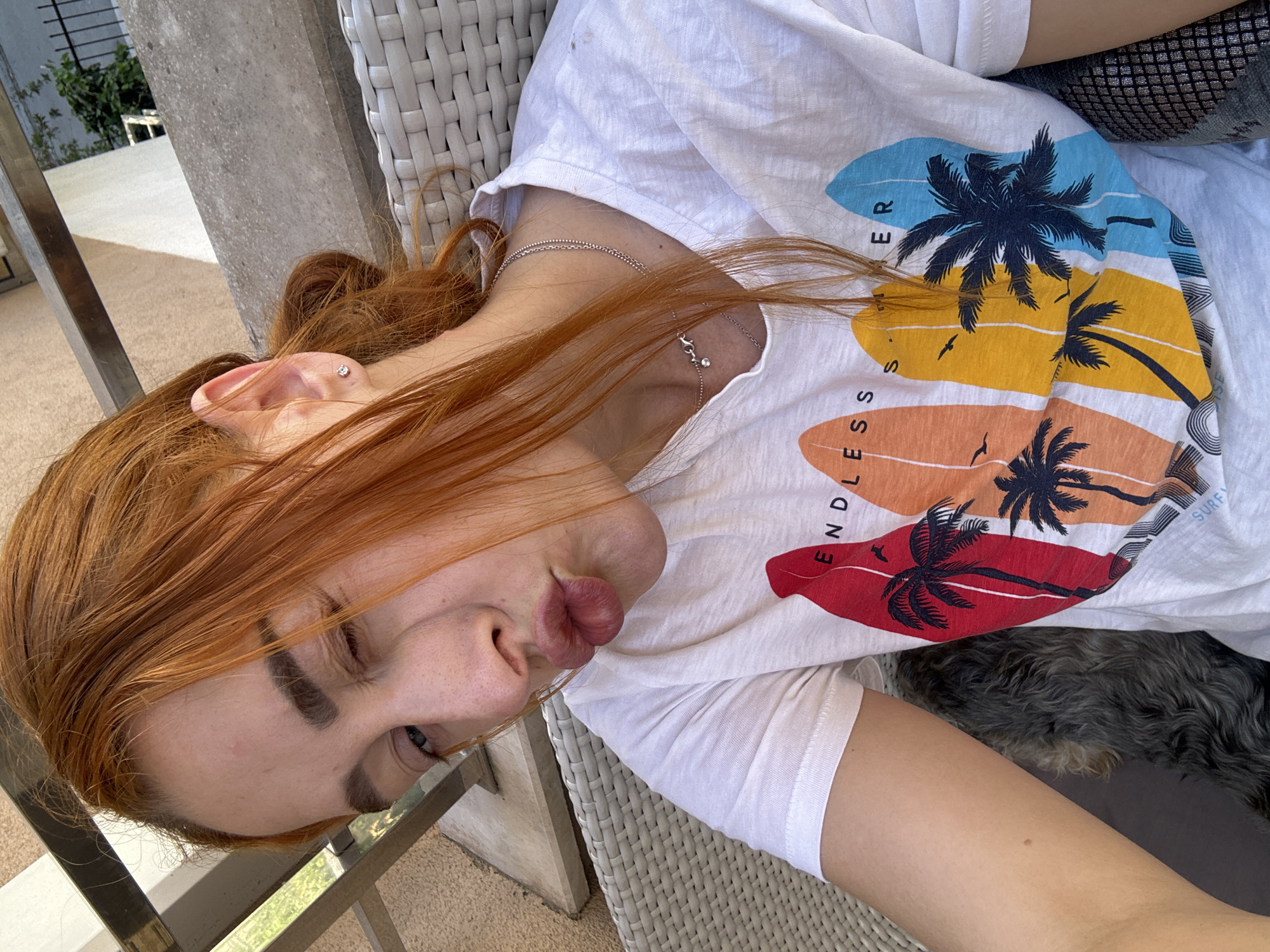 A woman with red hair is lying on a white wicker chair on a balcony or patio. She is wearing a white t-shirt with a tropical design featuring palm trees and the words "Endless Summer." The location appears to be in an outdoor space, possibly in the city of Budva, Montenegro, as suggested by the metadata.
